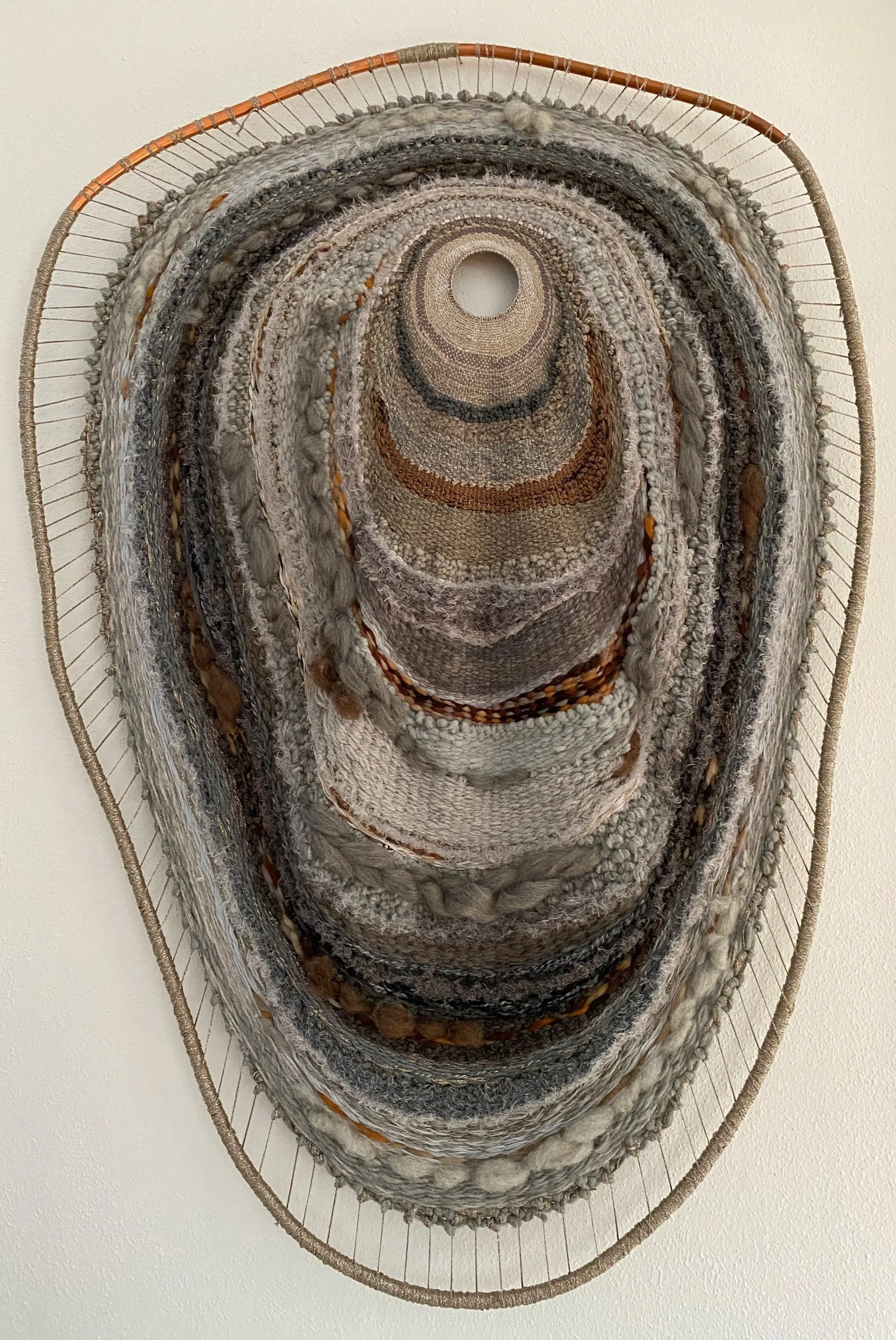 Oval wall art piece made of woven fabric and threads, featuring concentric shapes in earthy tones of beige, brown, and gray, mounted on a wall.