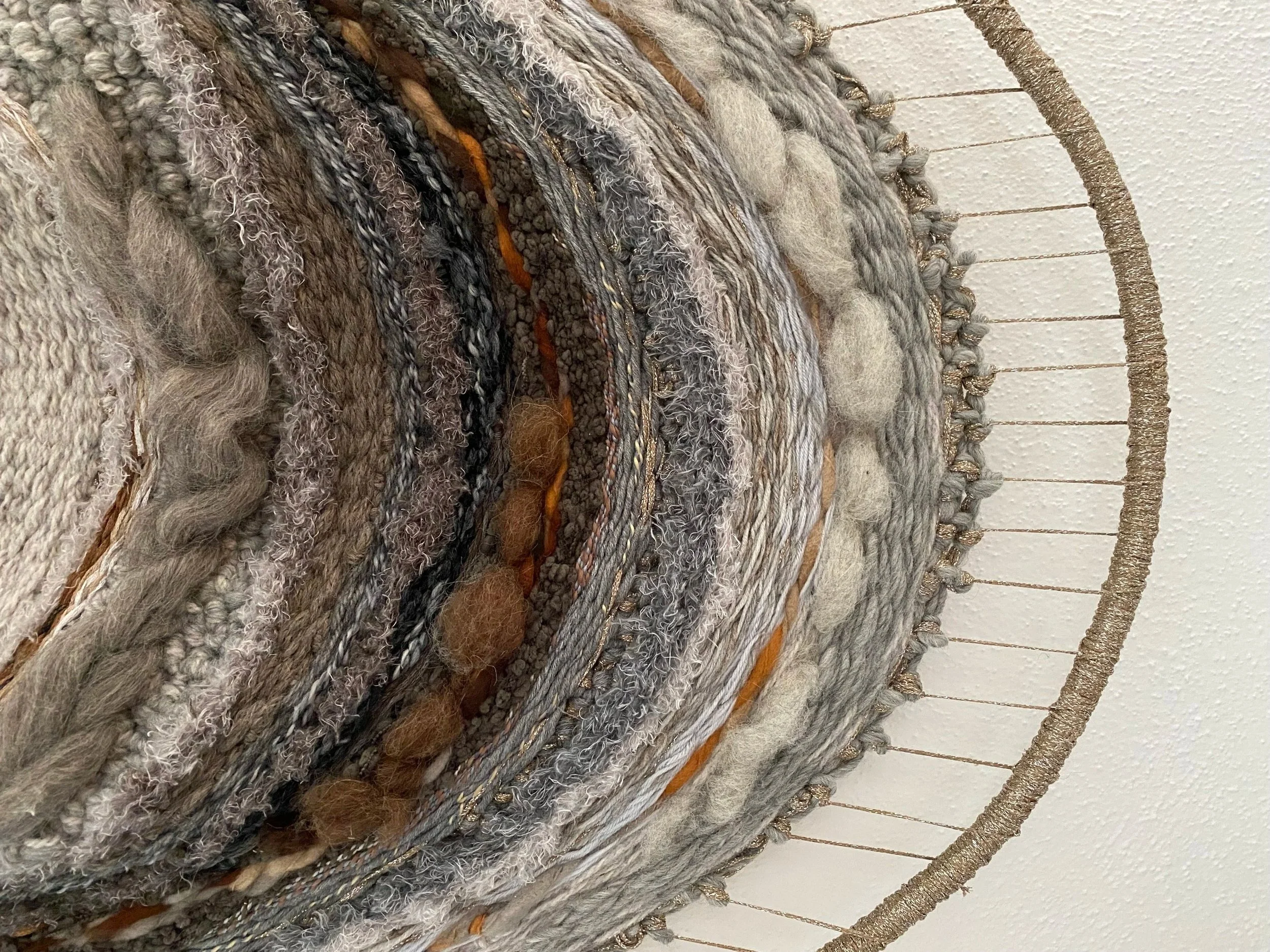Close-up view of a circular woven textile wall art with various textures and shades of neutral colors, mounted against a white ceiling.