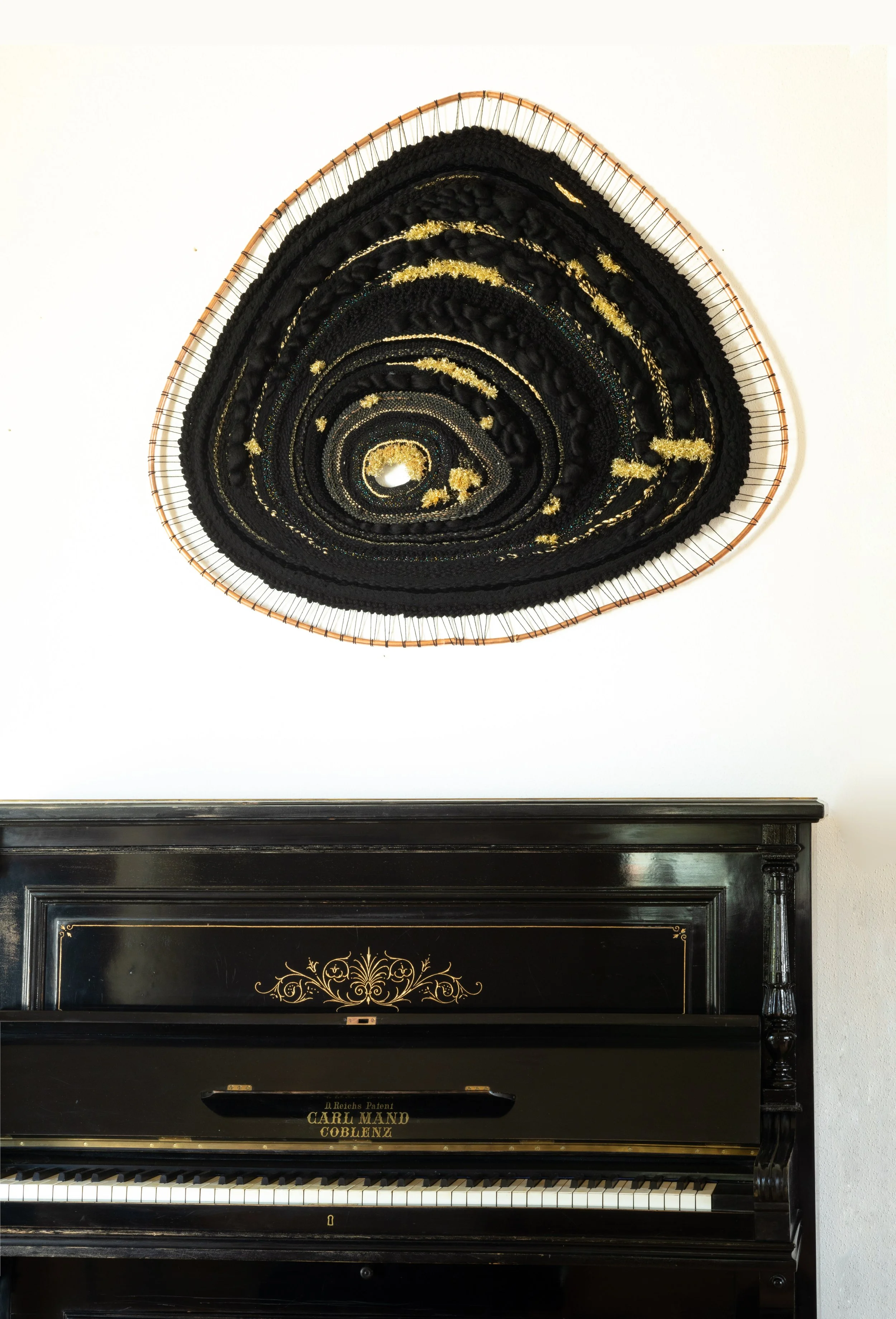 A black upright piano with gold decorative accents and the text "Carl Mand Coblenz" on the front, beneath a large oval-shaped artwork on the wall, which appears to be a woven or textile piece with black and gold patterns.