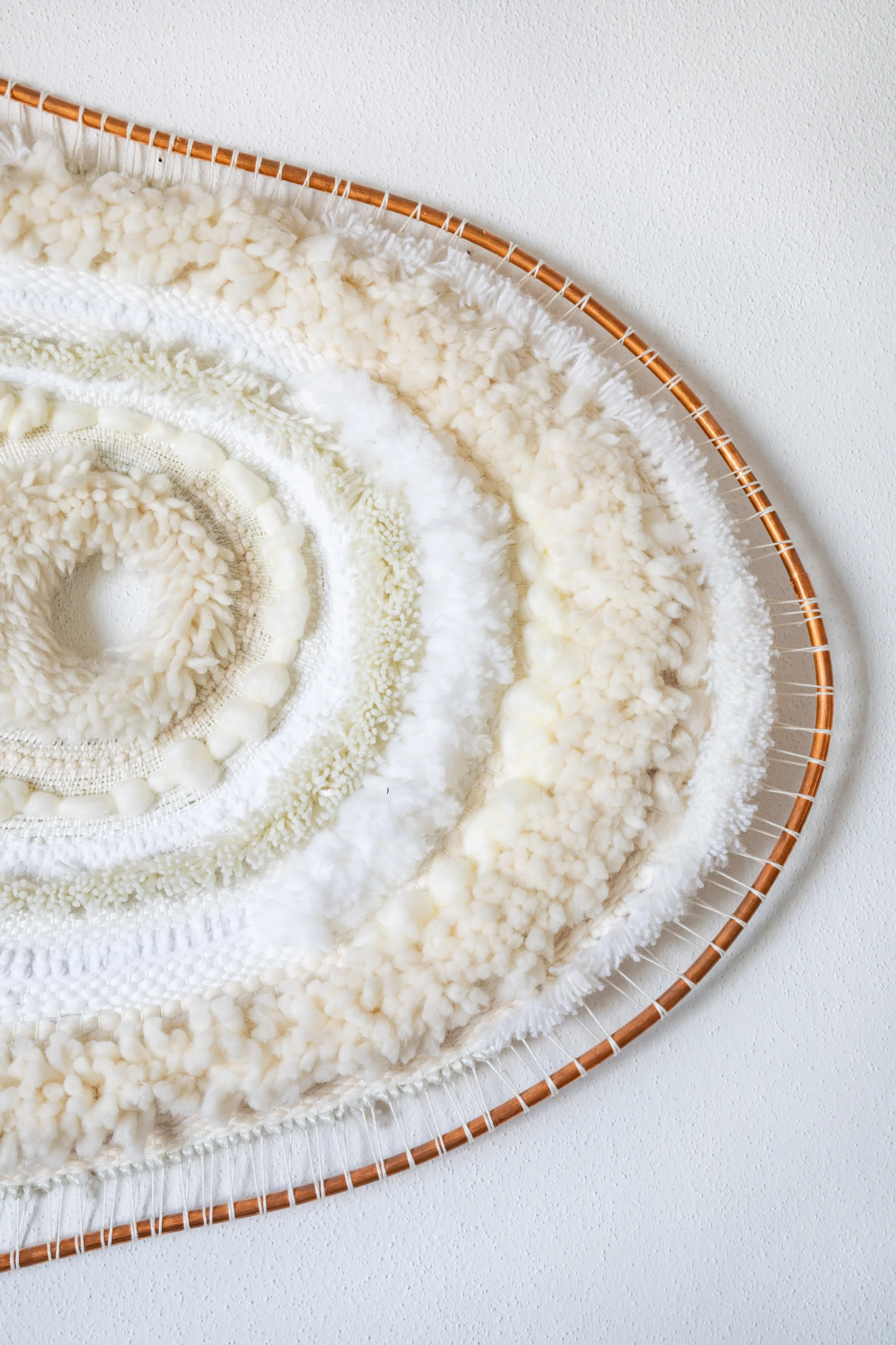 Close-up of a white, textured, and fluffy art installation or weaving, arranged in a spiral on a circular frame against a white wall.