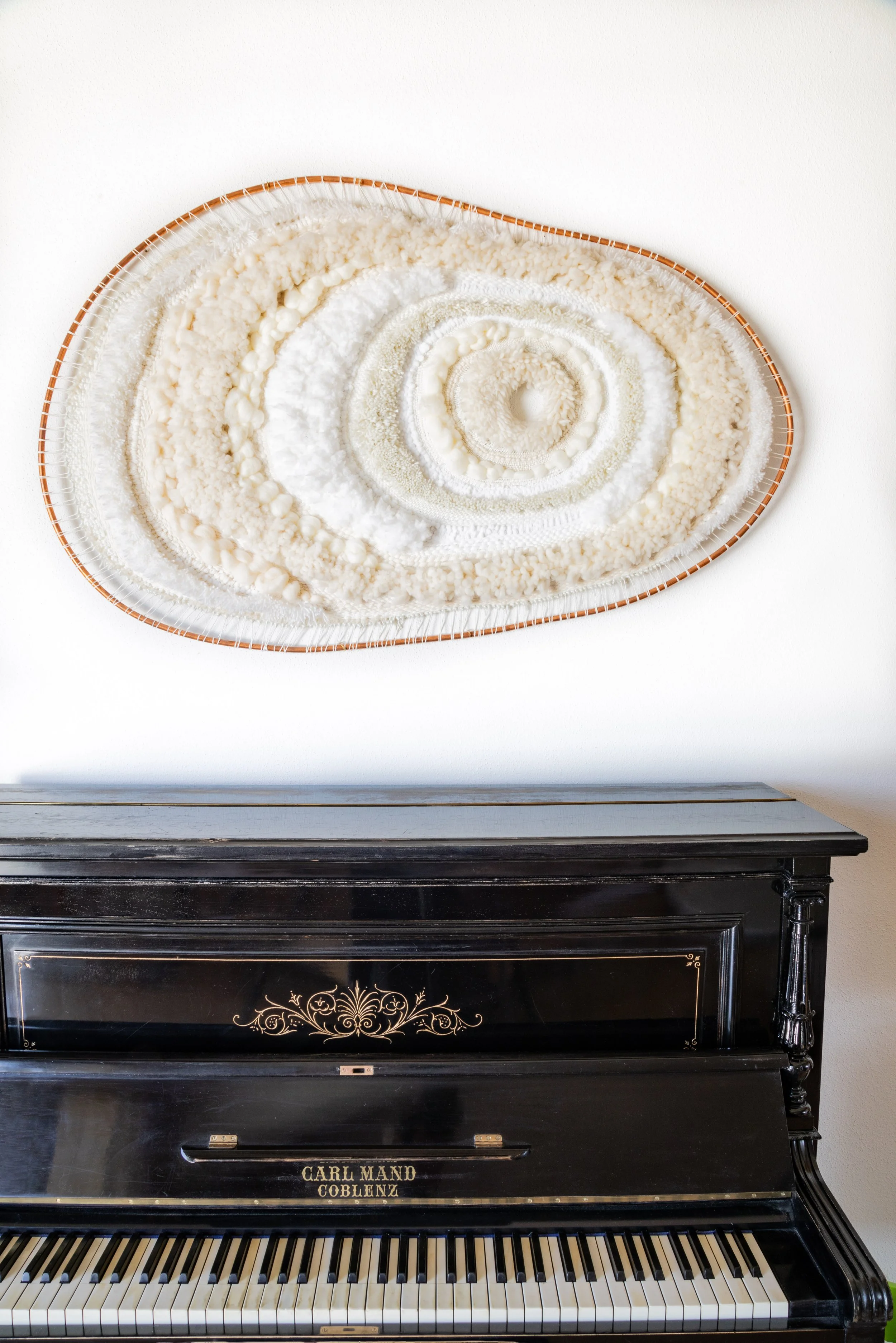 A black upright piano with gold decorative elements and white keys, positioned against a white wall, with an oval-shaped art piece above it that features a textured, circular, spiral pattern in shades of white and beige.