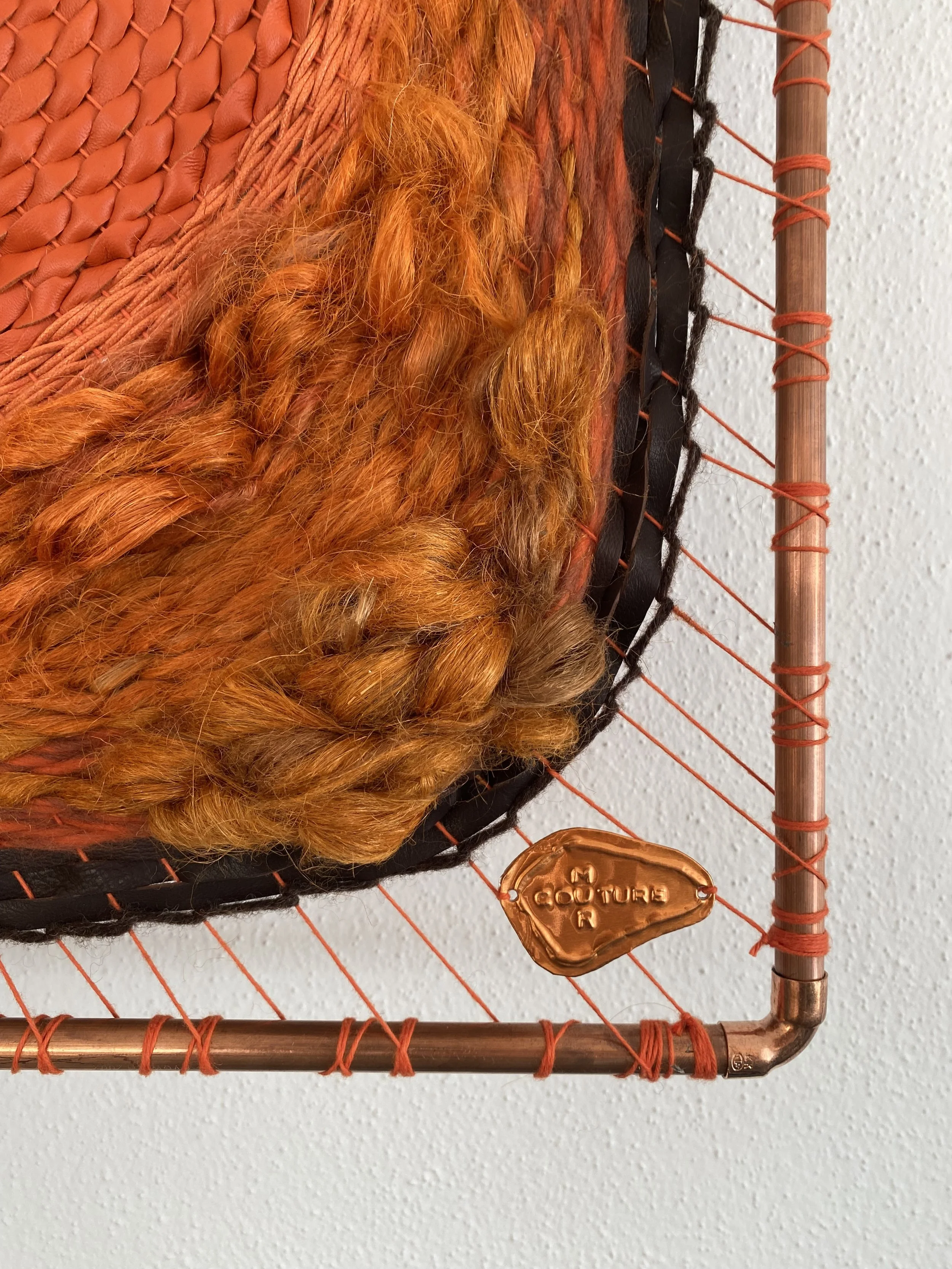 Close-up of a decorative wall art piece made of woven copper wire, with orange and black yarn, a spray of orange hair, and a small copper tag that says 'COUTURE'.
