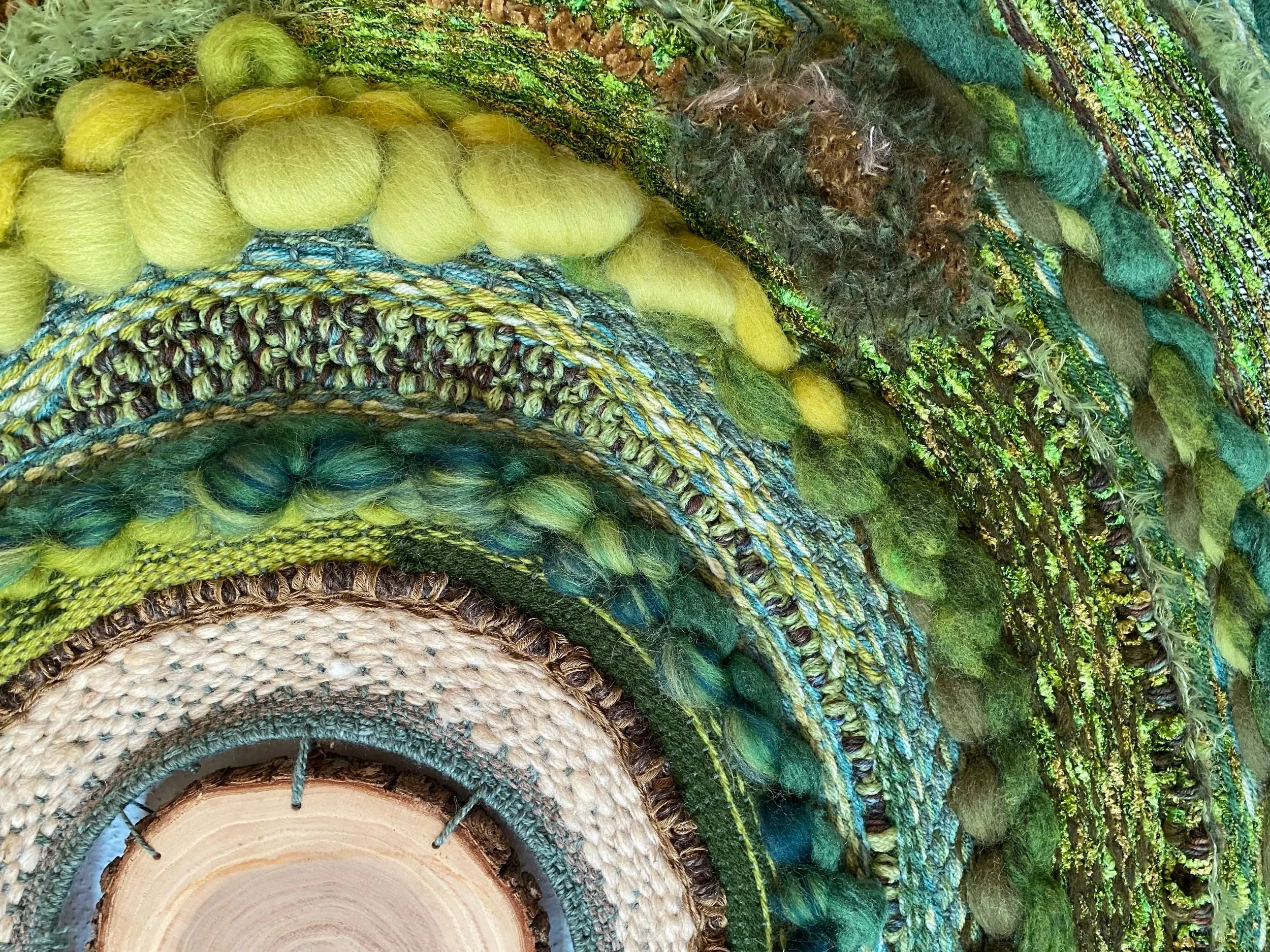 Close-up of a fiber art piece made of various textured yarns in shades of green, blue, and beige, arranged in concentric circles.