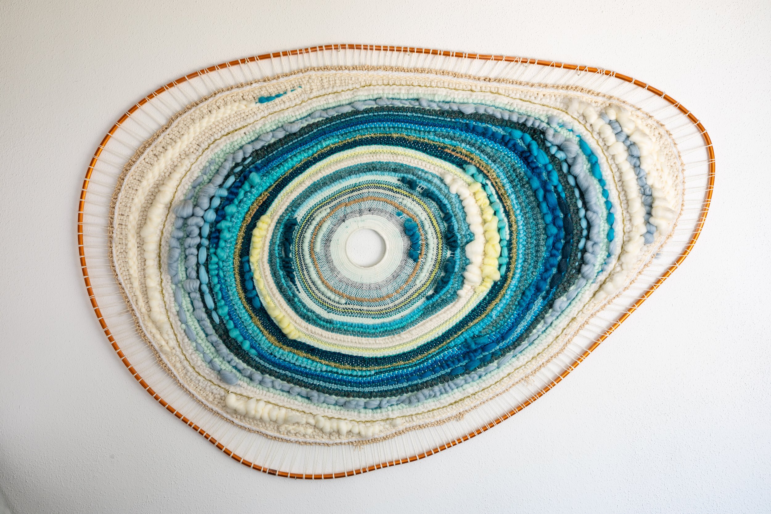 A colorful circular woven wall art with shades of blue, white, and yellow, mounted on a white wall.