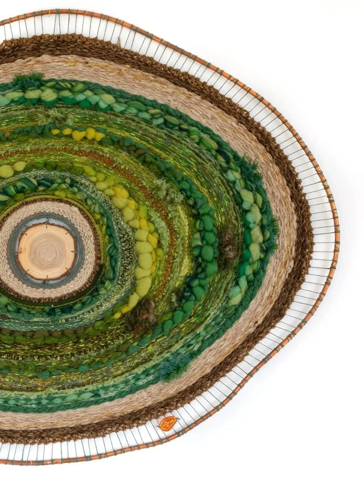 A circular woven wall art piece made from various textured and colored yarns, fabrics, and natural materials, mounted on a wooden base with a metal wire frame.