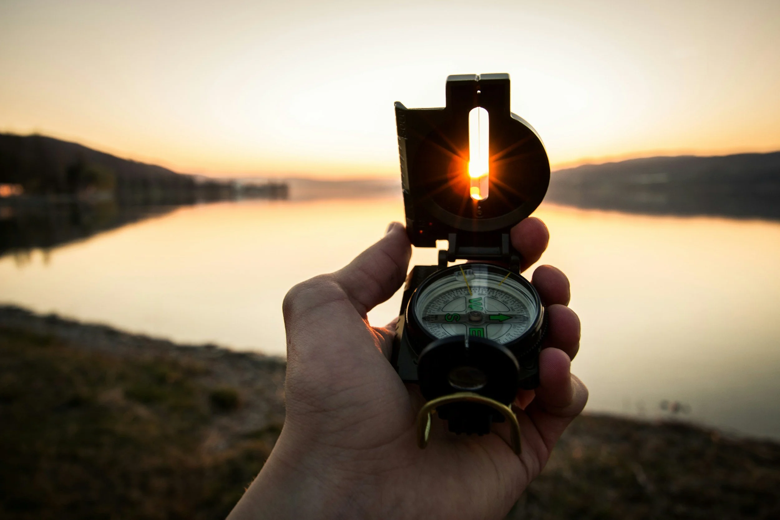 A person holding a compass with a sunset over a lake in the background.