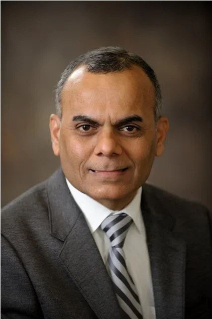 A professional headshot of a middle-aged man with short, dark hair, wearing a gray suit, white shirt, and striped tie, against a brown blurred background.