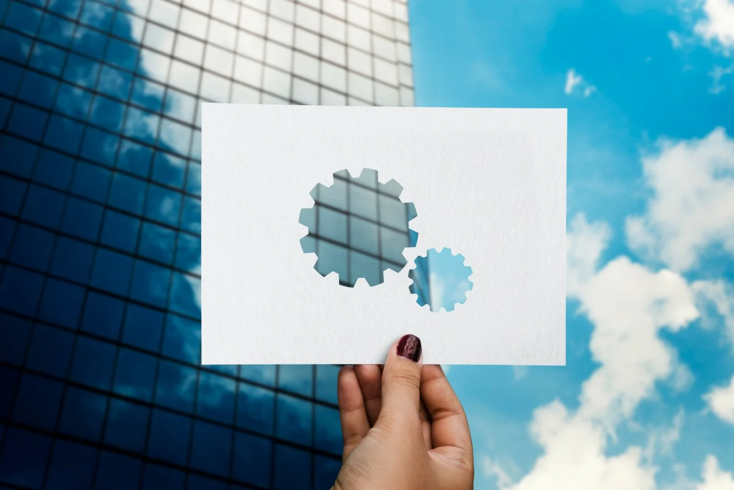 Person holding a white card with cutouts of gears against a modern glass building and blue sky with clouds.