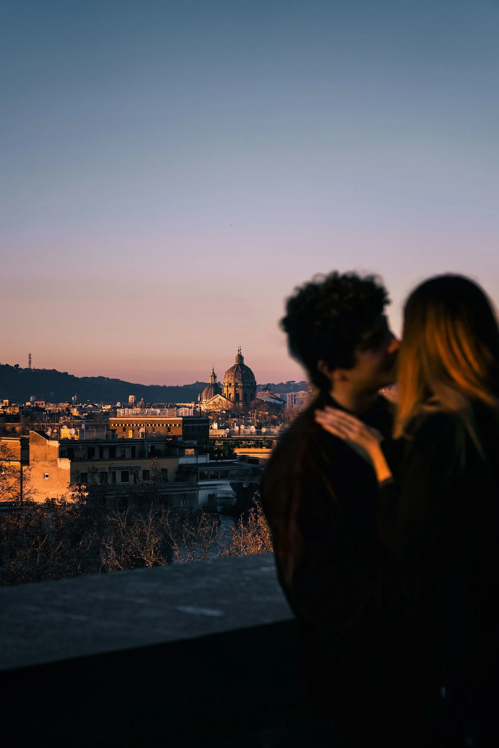 Una pareja besándose en primer plano con la ciudad y una cúpula de Roma en el fondo al atardecer