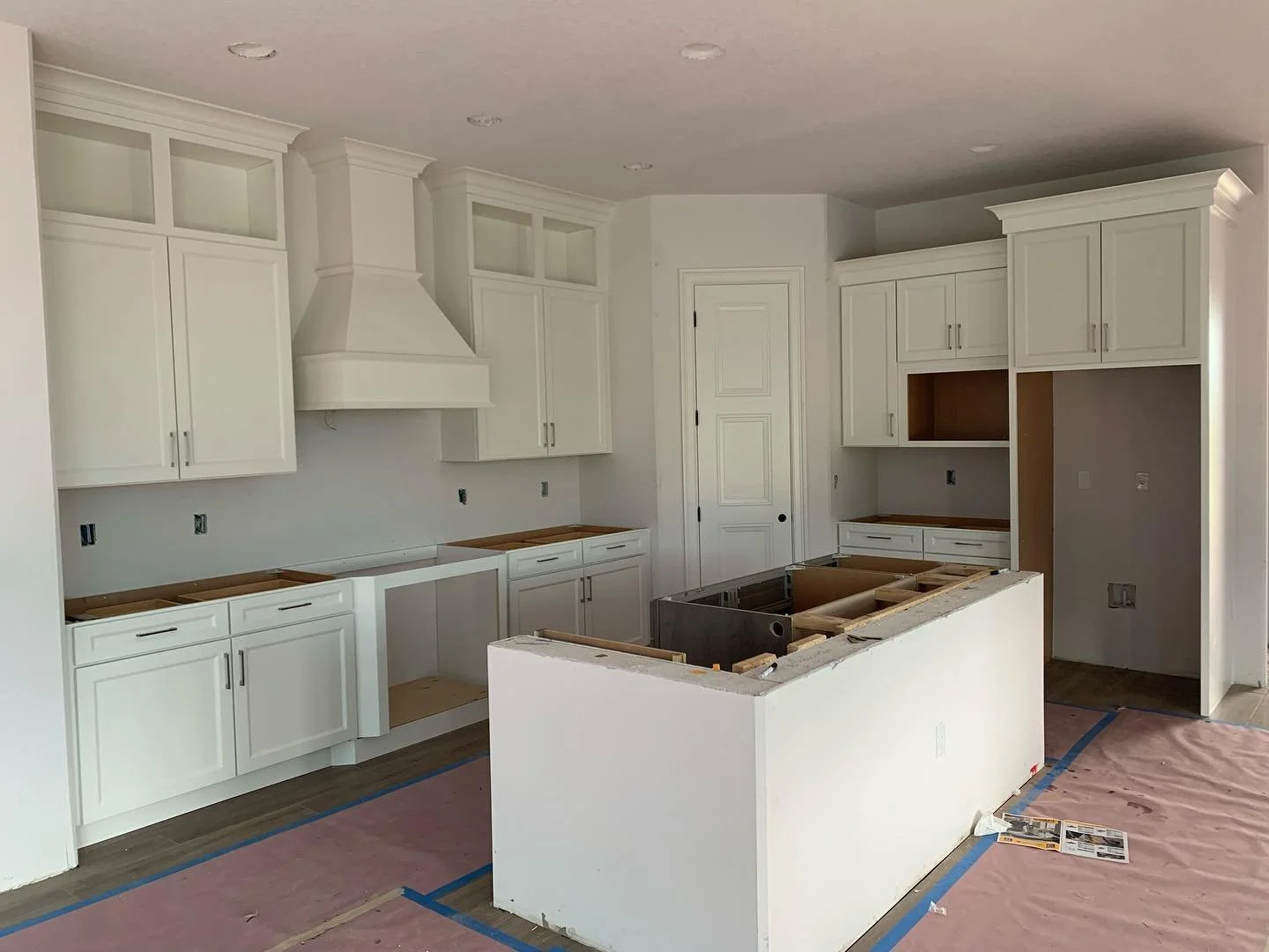 Kitchen under construction with white cabinets and a large island, no countertops installed yet, construction materials on the floor.