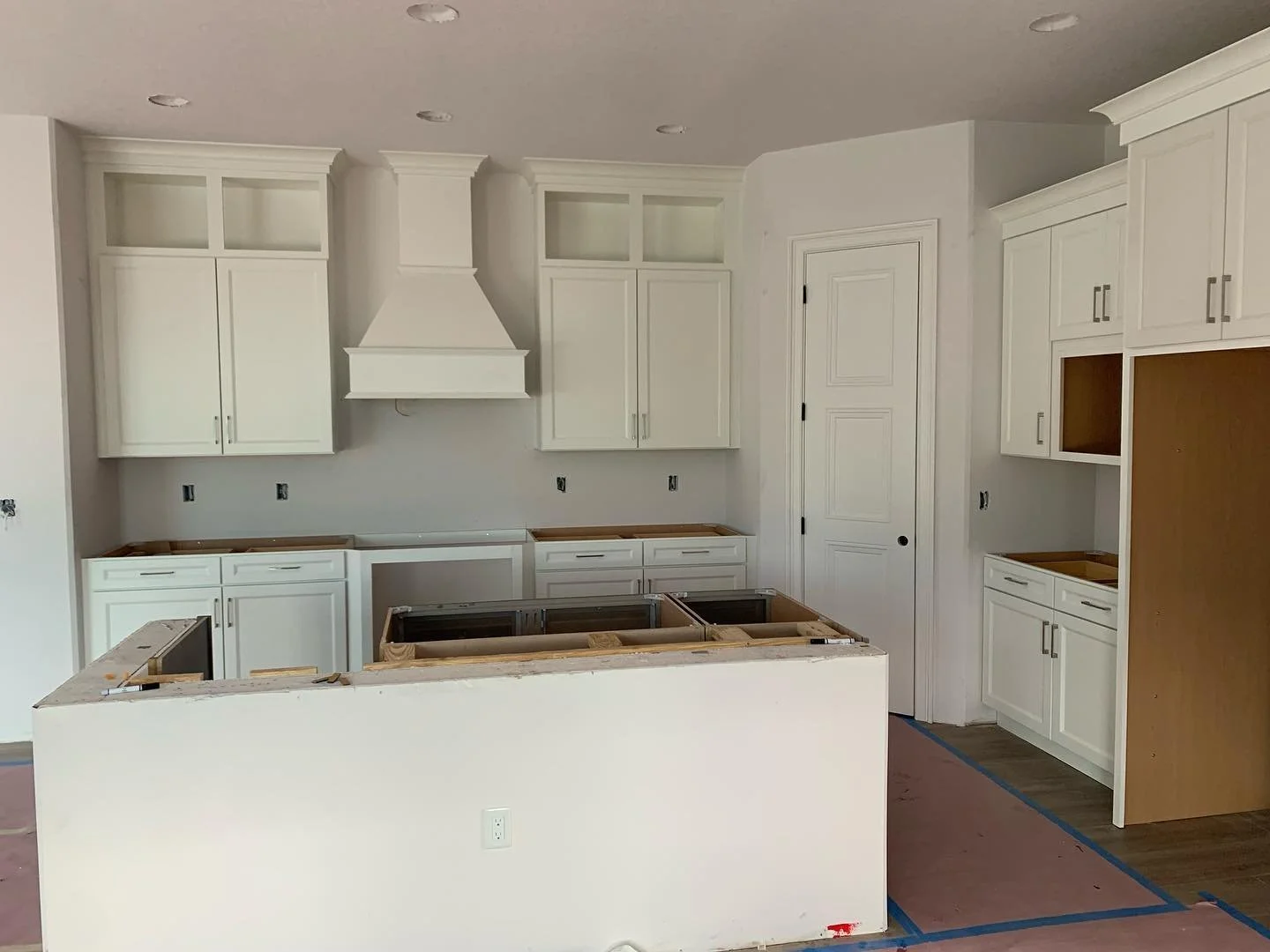 Kitchen under construction with white cabinets, a white range hood, and an island frame, no countertops installed yet.