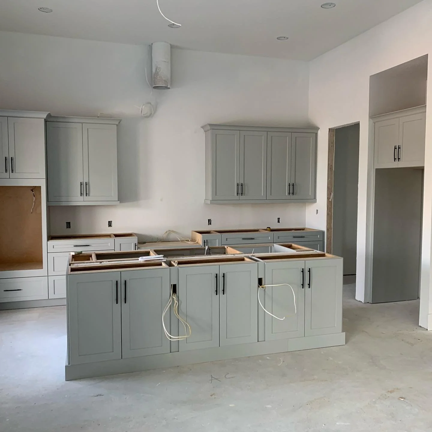 Kitchen under construction with grey cabinets and open island space, no appliances installed, unfinished walls and flooring.