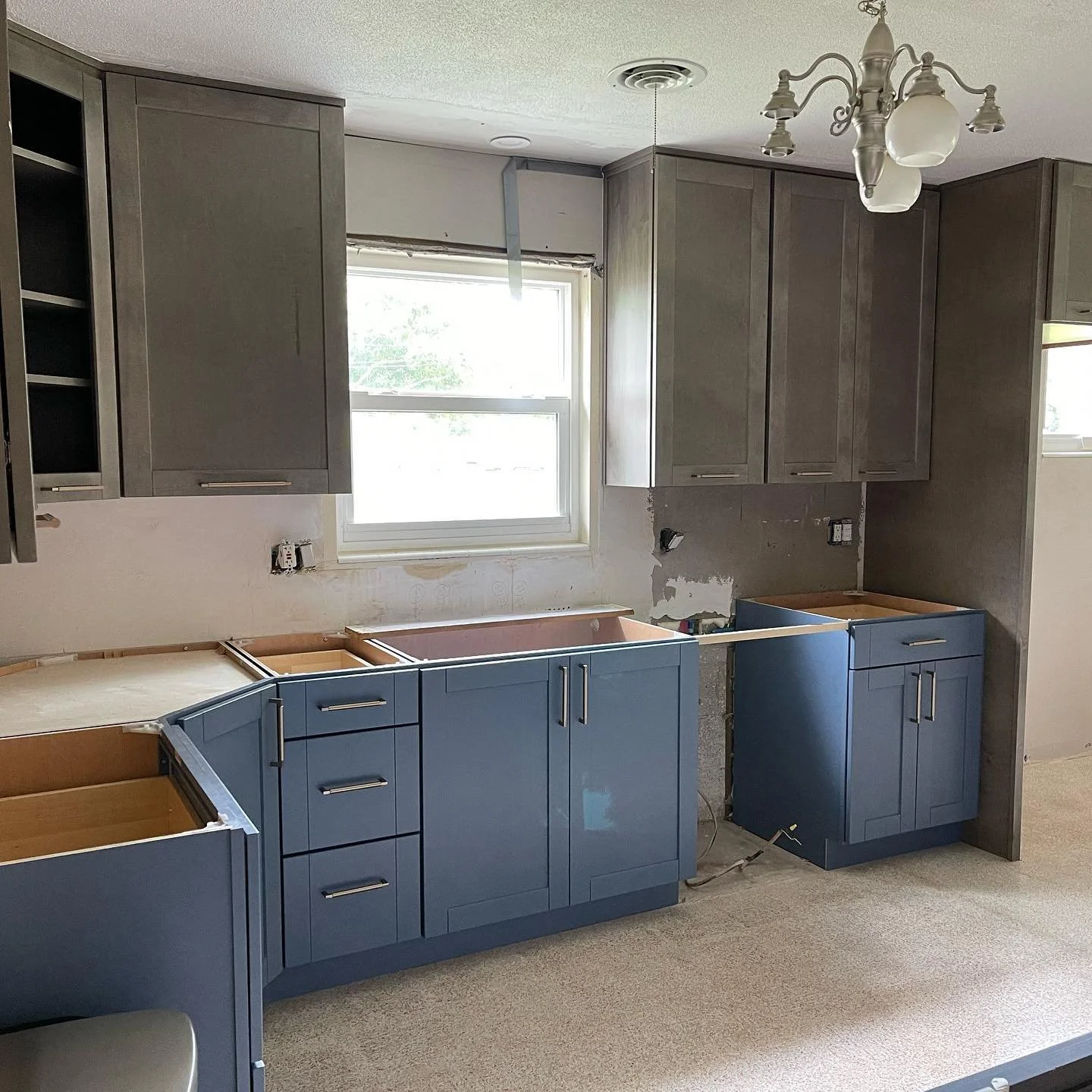 Kitchen cabinets being installed with blue lower cabinets and gray upper cabinets, with a window and a ceiling light fixture.