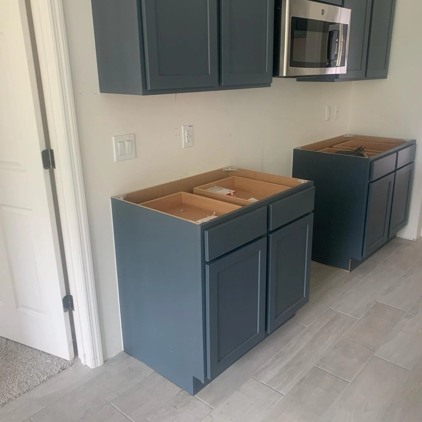 Two unfinished kitchen base cabinets with dark blue paint in a room with tiled floor, wall outlets, a microwave clock, and upper cabinets mounted on the wall.