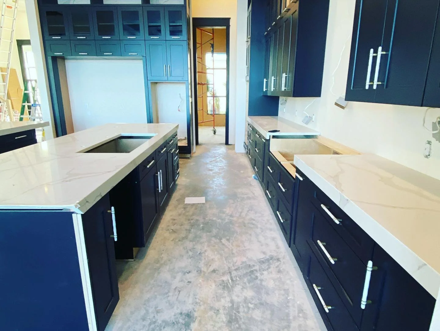 Kitchen under construction with navy blue cabinets, white countertops, and open space for appliances, with visible construction tools and scaffolding outside the window.