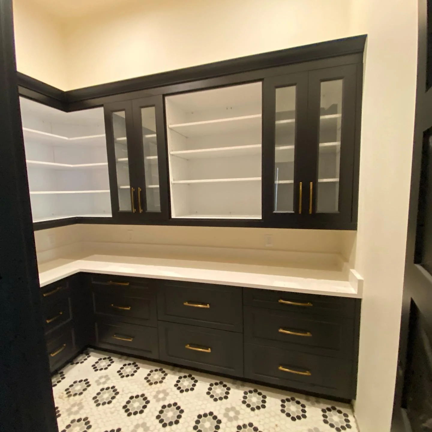 Black kitchen cabinets with gold handles and white countertop, featuring glass doors on upper cabinets and white shelves inside, with black and white patterned floor tiles.