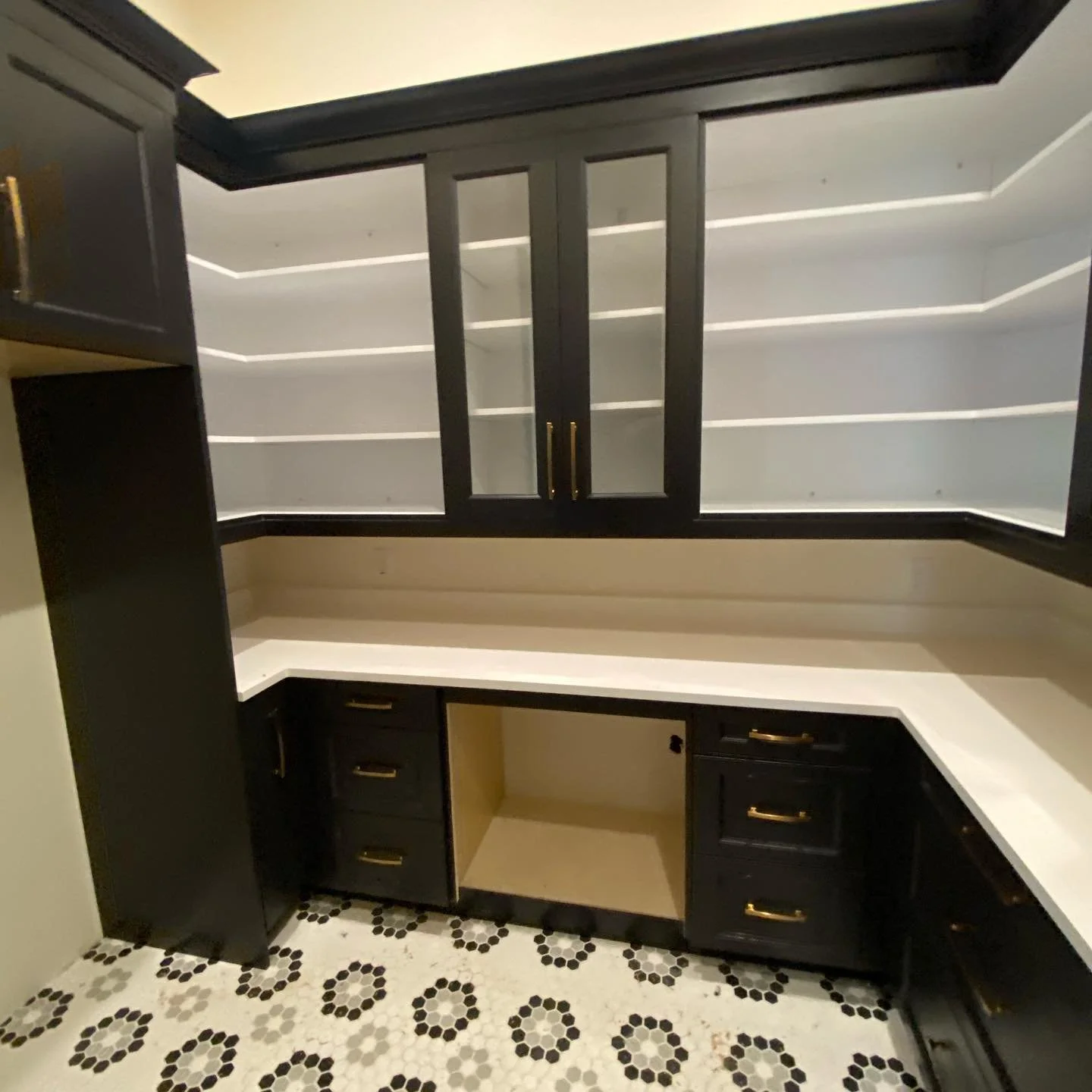 Empty kitchen cabinet with black cabinetry, gold handles, white countertops, some shelves behind glass doors, and a black-and-white patterned floor.