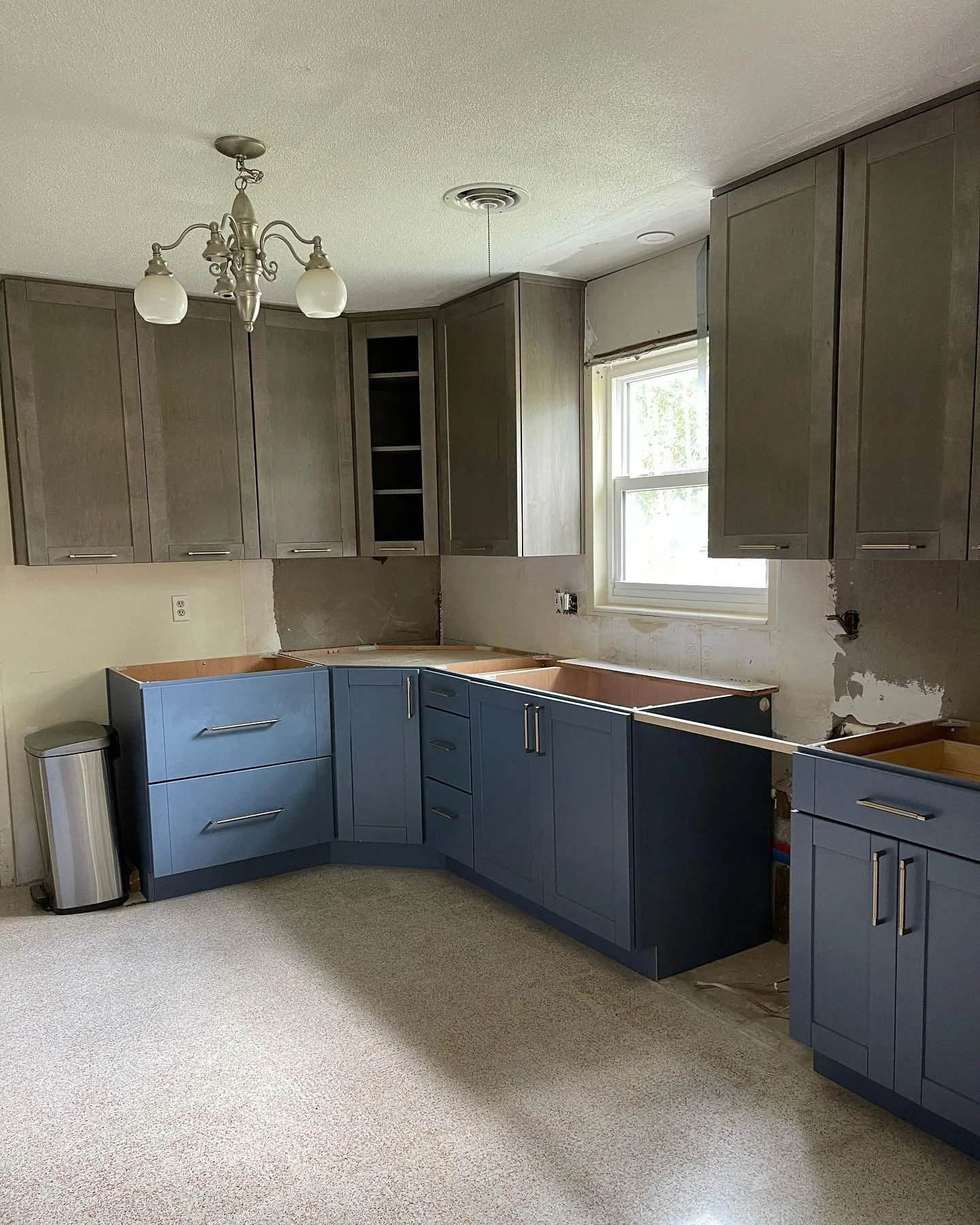 Kitchen under renovation with blue lower cabinets, gray upper cabinets, a window, and a chandelier, with some areas unfinished.