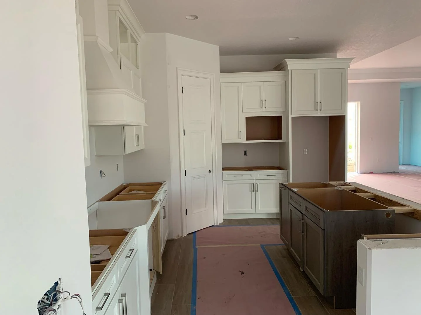 Kitchen under construction with white and dark cabinetry, countertops not yet installed, and protective pink flooring covering the floor and island.