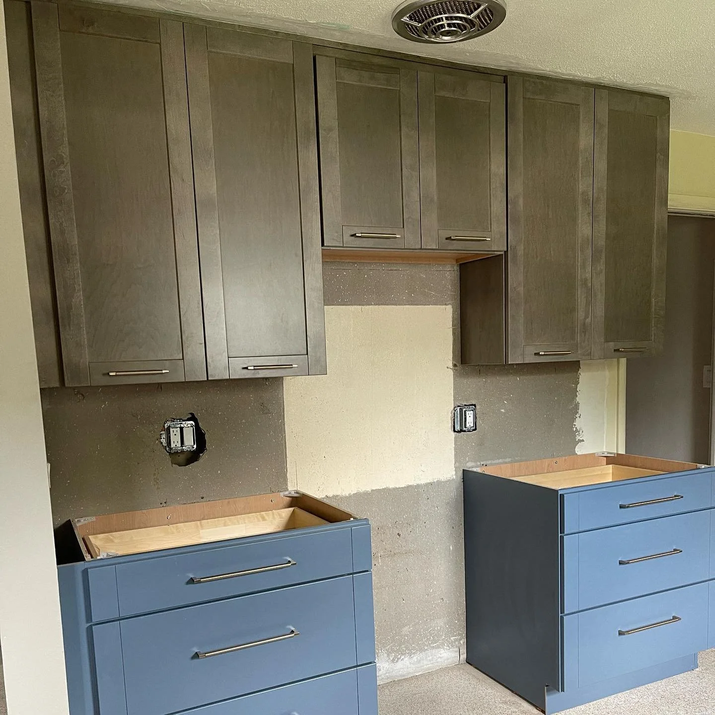Kitchen cabinets under installation with upper gray cabinets and lower blue drawers, electrical outlets visible and a partially finished wall.