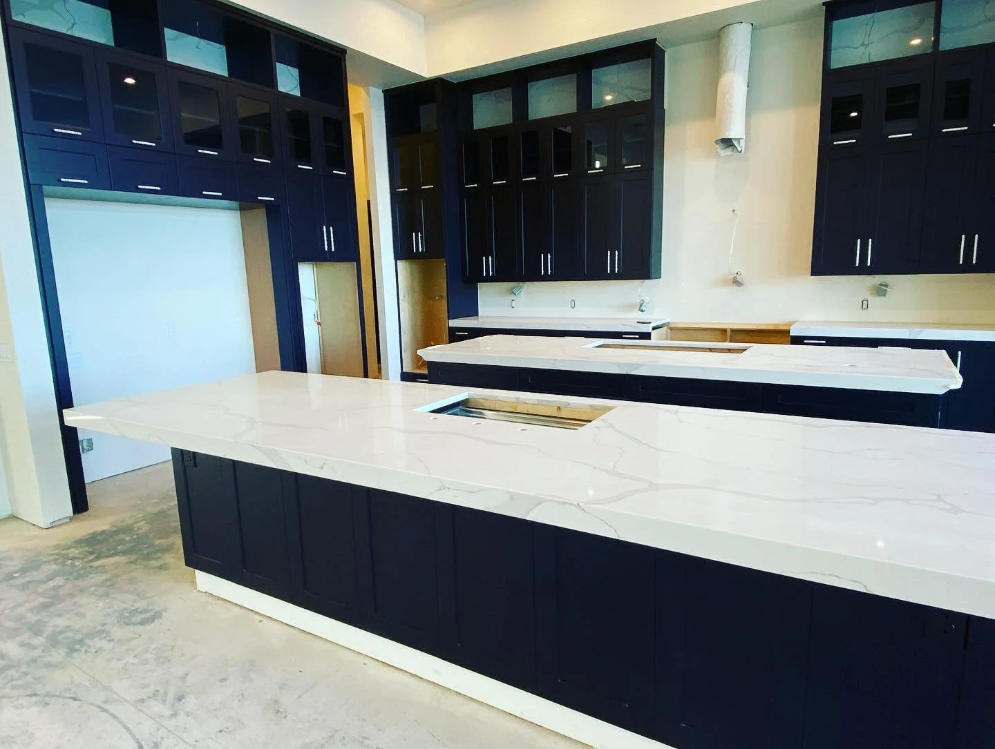 A modern kitchen under construction with black cabinets, white marble countertops, and no appliances installed yet.