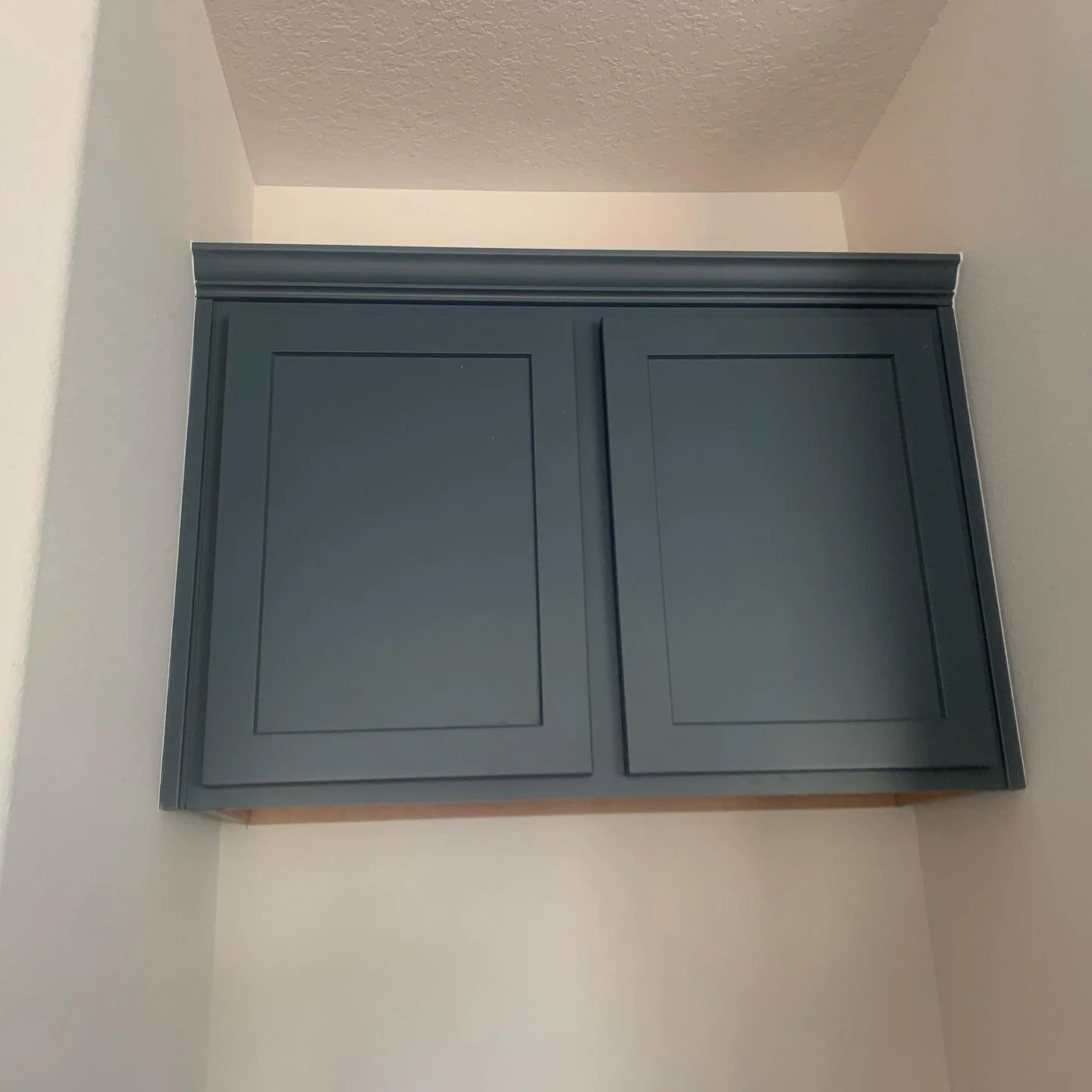 Dark gray cabinet mounted on a wall in a corner of a room with white walls and a textured ceiling.