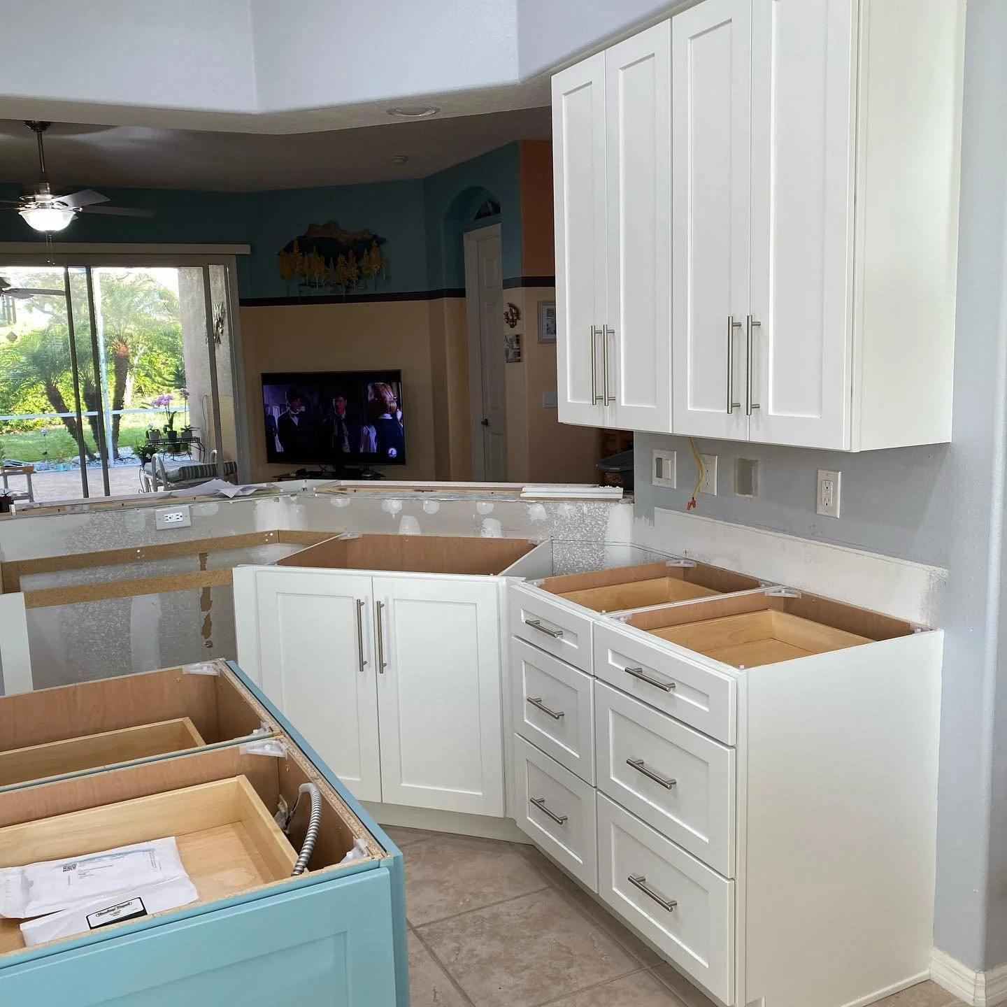 Kitchen cabinets installation in progress with white cabinets and no countertops, new drawers and cabinet doors visible, and a view into the living room with a TV and sliding glass door leading outside.
