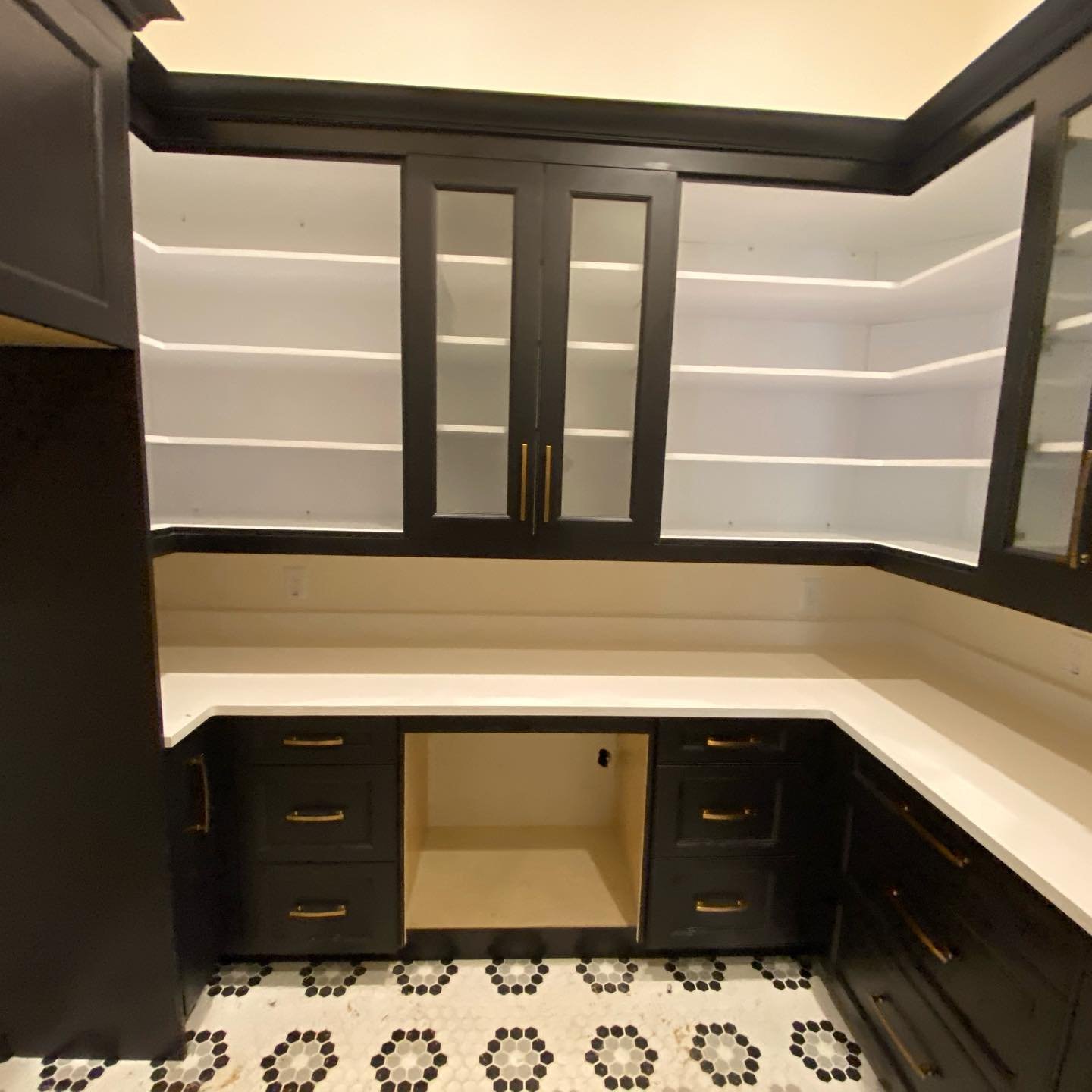 Empty black kitchen cabinets with glass doors and open shelves, white countertops, black and white patterned tile floor.