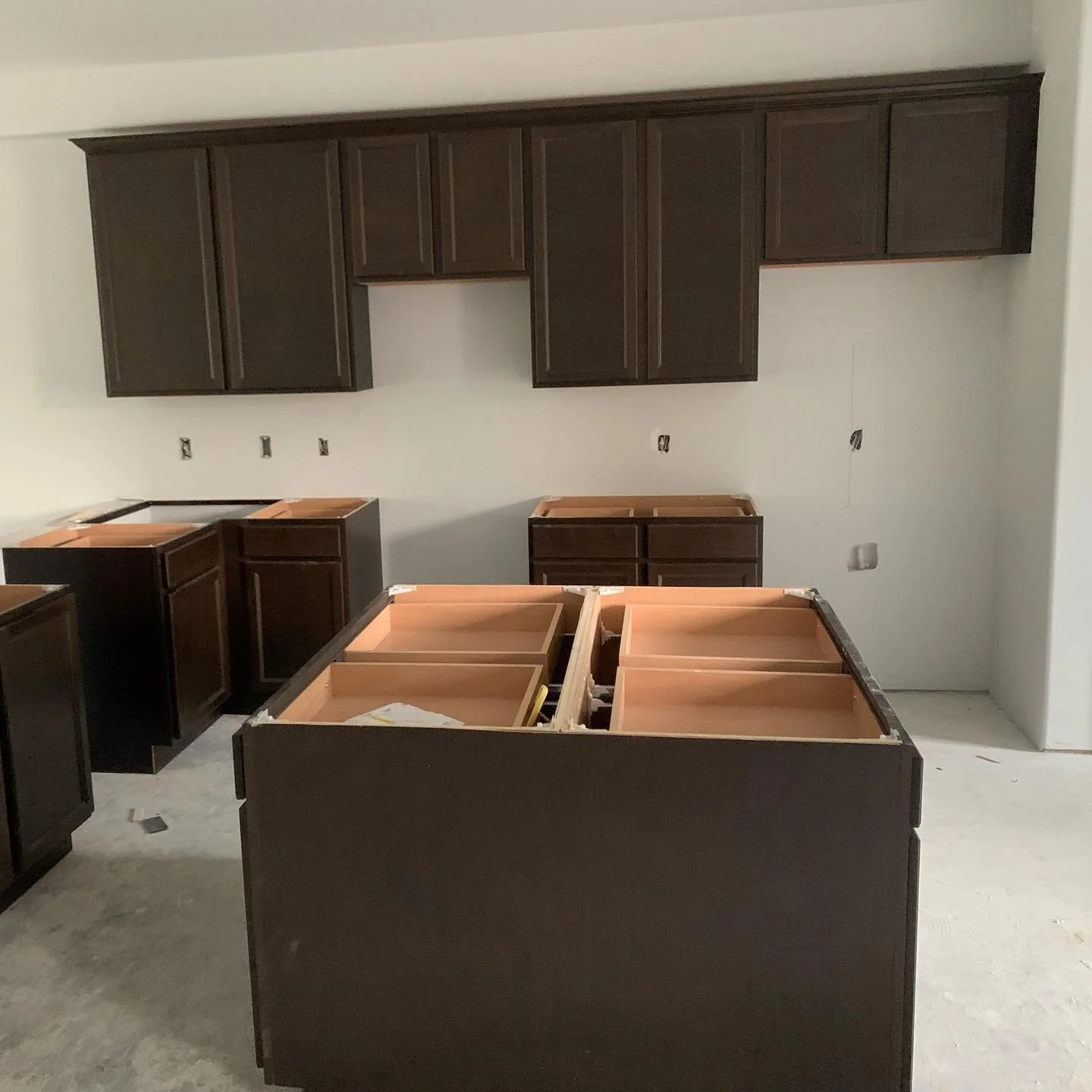 Kitchen under construction with dark wooden cabinets and an island. The upper cabinets are mounted on the wall, and the lower cabinets are partially installed. The countertop is not yet in place.