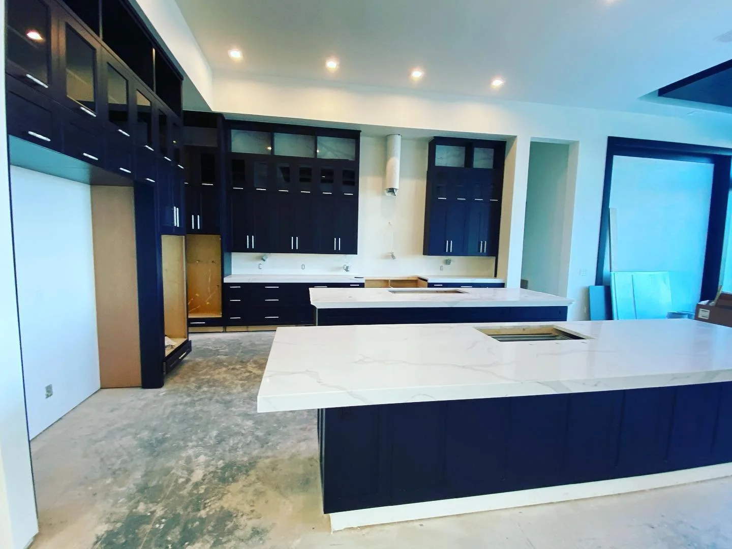 Modern kitchen under construction with dark wood cabinets, white marble countertops, and an island, with unfinished flooring and construction materials.
