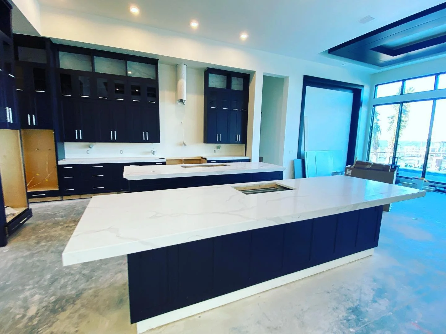 Modern kitchen with white marble countertops, dark blue cabinets, and large windows showing outside view. Unfinished cabinetry and construction materials present.