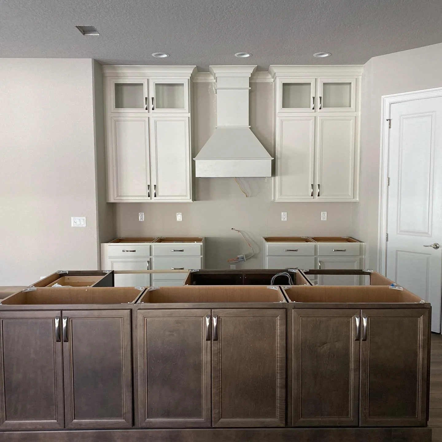 Kitchen cabinet installation in progress, with brown lower cabinets and white upper cabinets and range hood, no countertops yet.