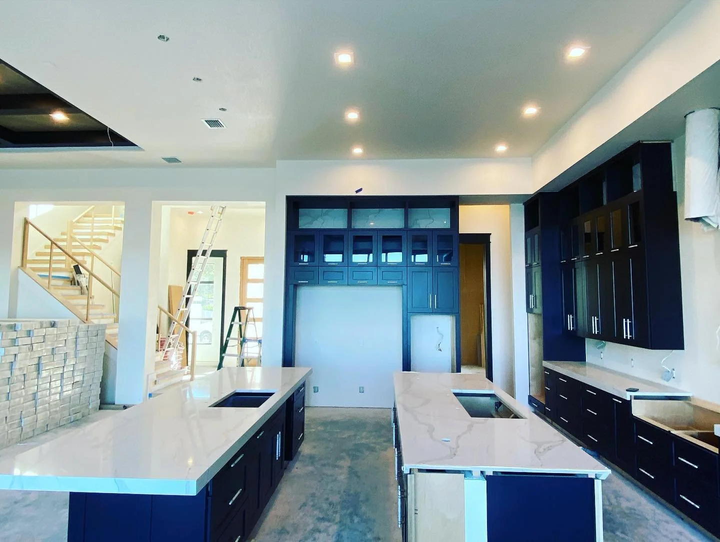 Interior of a kitchen under construction with black cabinetry, white countertops, and two kitchen islands, one with a cooktop, in a modern home with exposed concrete flooring and unfinished walls.