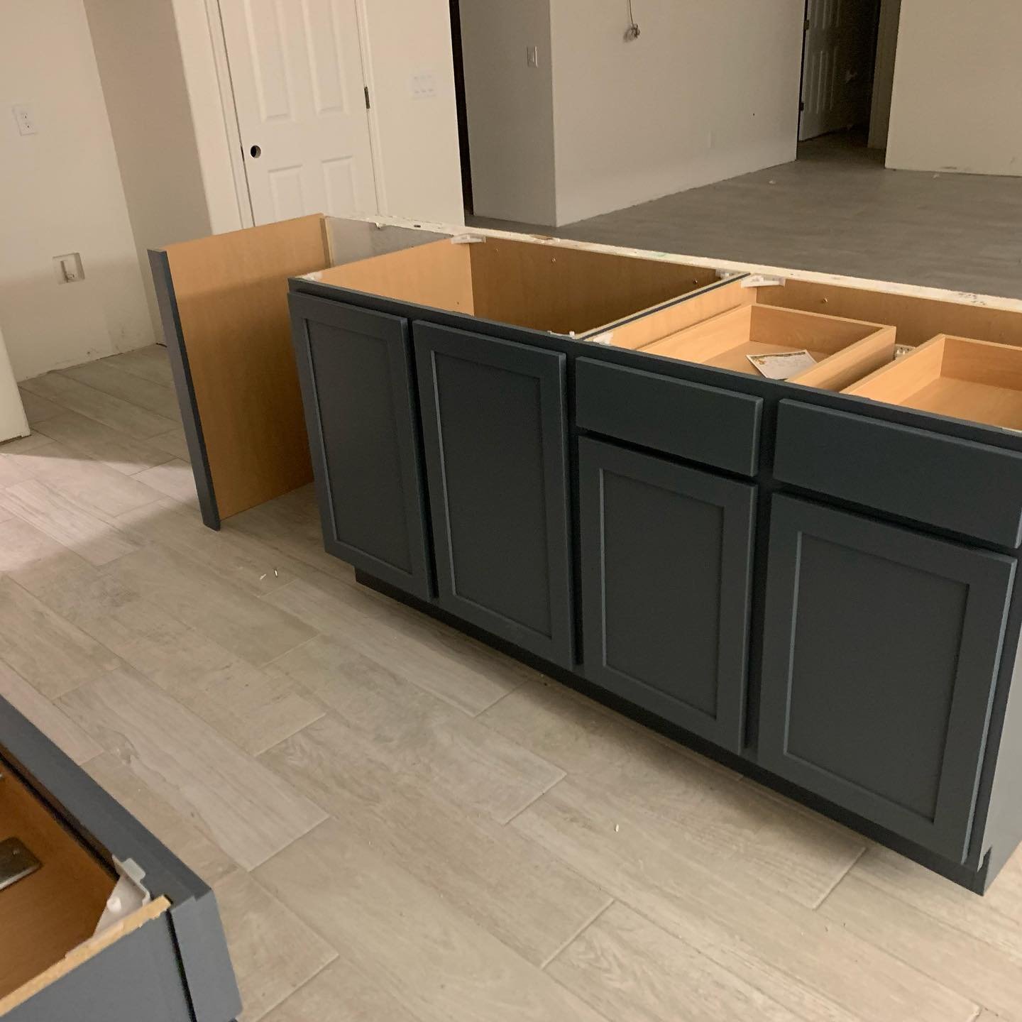 Partially installed kitchen cabinets in a kitchen under renovation. The lower cabinets are painted dark gray with recessed panel doors, and the countertop is missing, revealing the wooden base. The floor is covered with light-colored wood-look tiles.