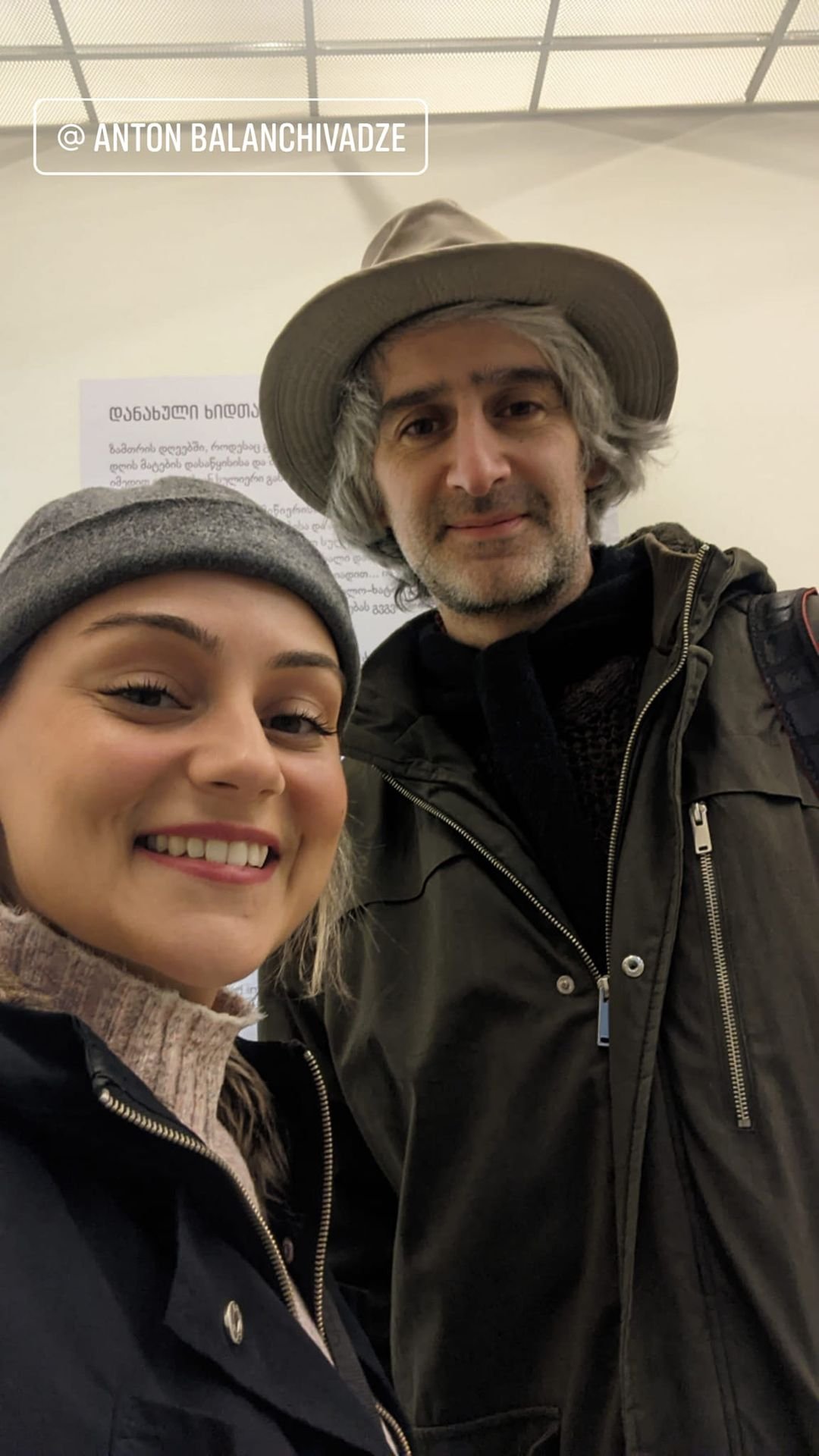 Two people smiling for a selfie indoors. The woman on the left has light skin, blonde hair, and is wearing a gray beanie, a beige sweater, and a black jacket. The man on the right has light skin, gray hair, and is wearing a light-colored hat and a da