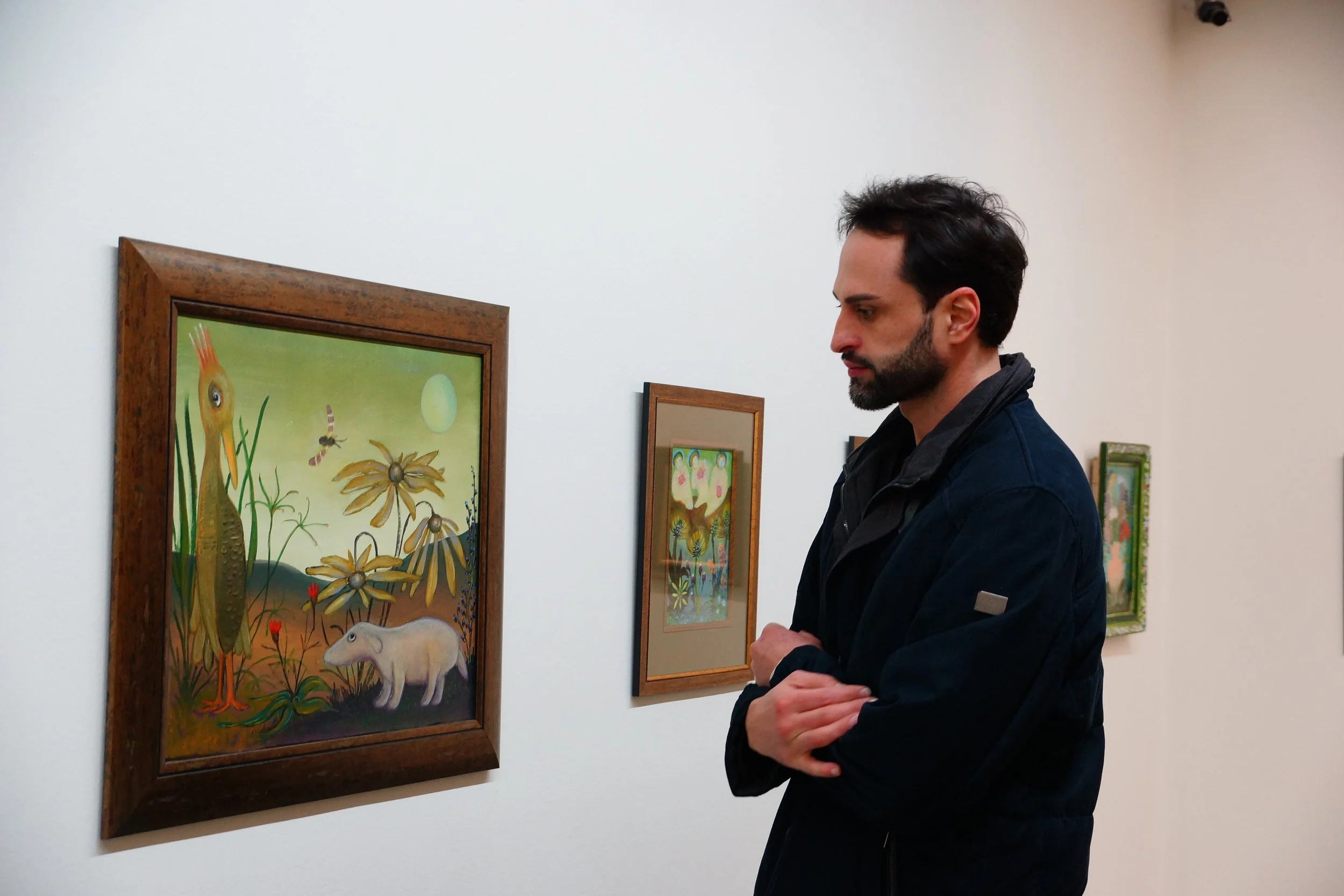 A man with dark hair, a beard, and wearing a dark jacket is standing in an art gallery, observing a colorful painting of a bird, flowers, a rodent, and insects on a white wall.