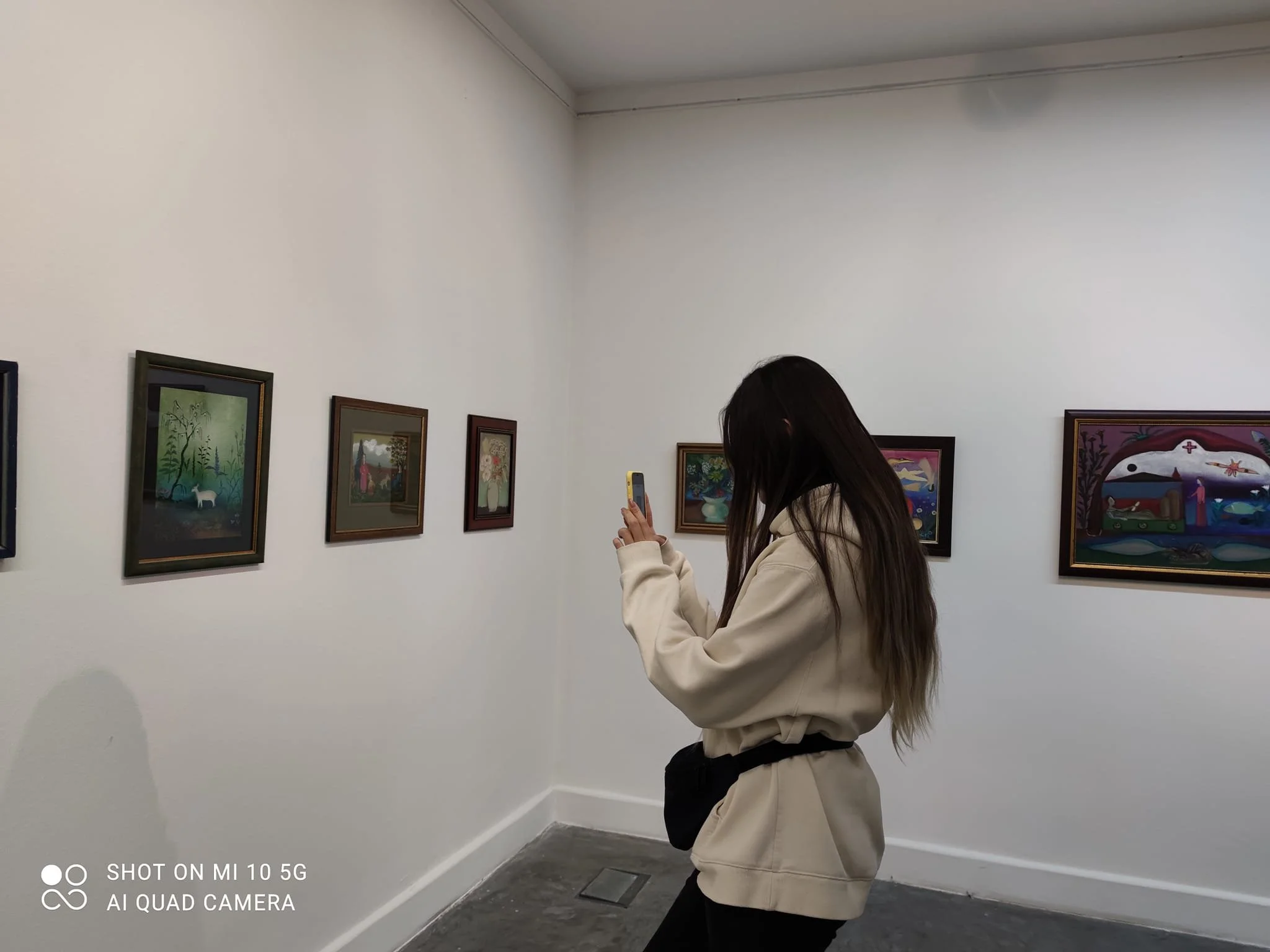 A woman with long dark hair, wearing a beige hoodie and black pants, taking a photo with her smartphone in an art gallery with framed paintings on white walls.