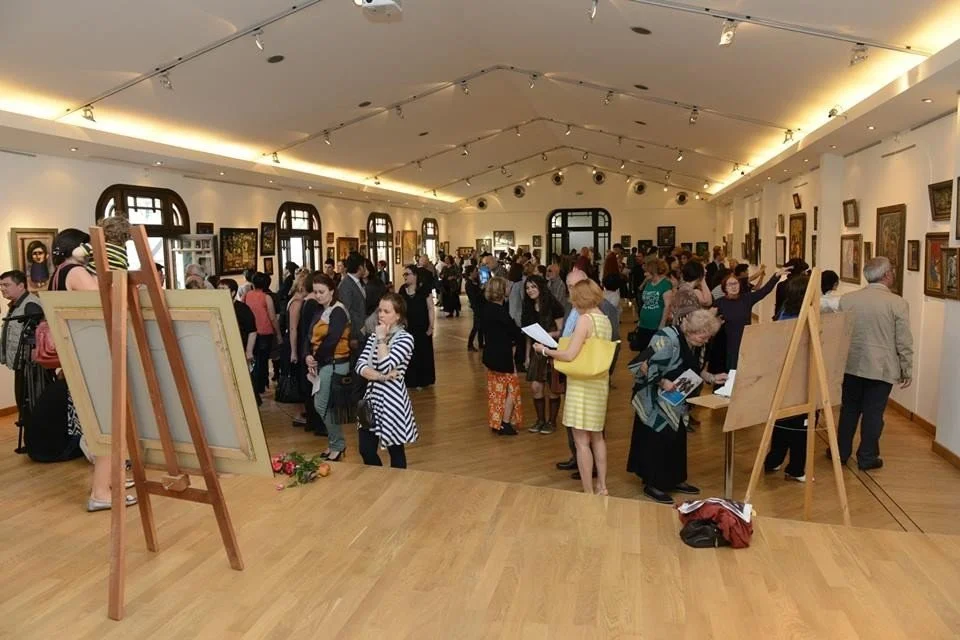 People viewing artwork in a spacious art gallery with paintings on the walls and large windows, some reading and others discussing.