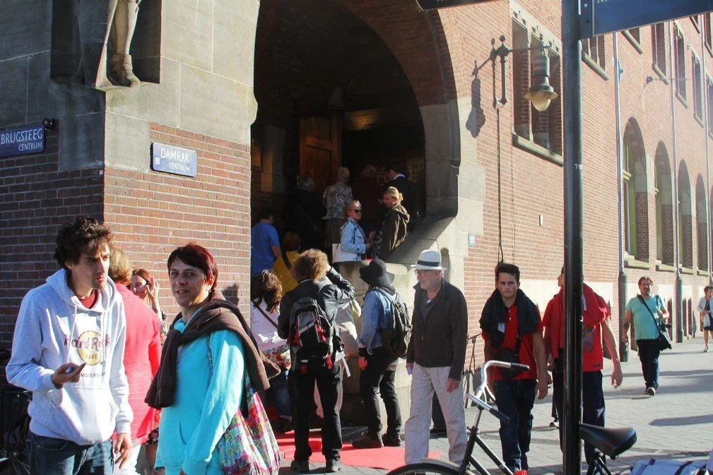 People waiting in line outside a building entrance on a city street, some engaged in conversation, with brick architecture and street lamps visible.