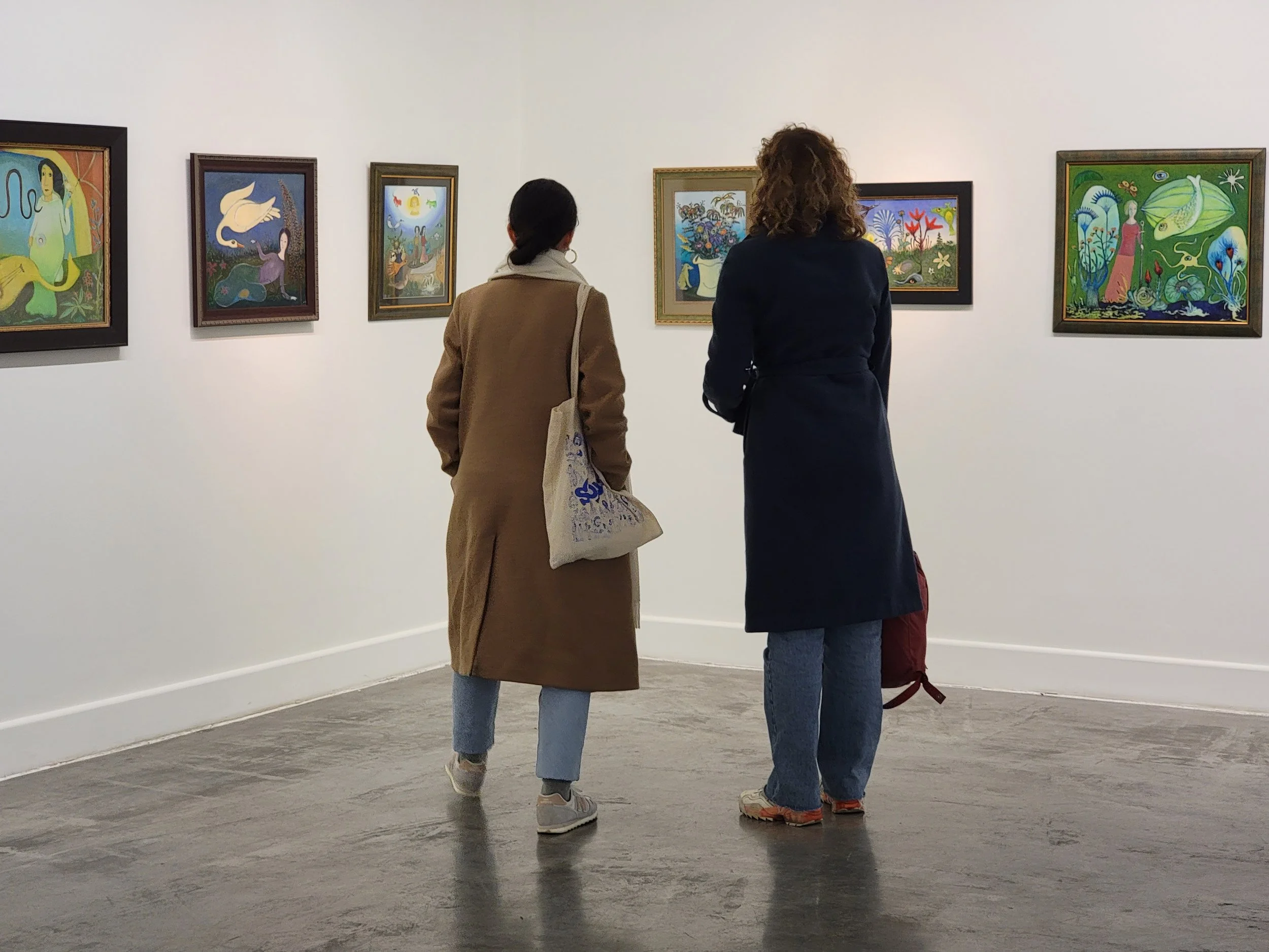 Two people stand in front of colorful paintings displayed on a white gallery wall.