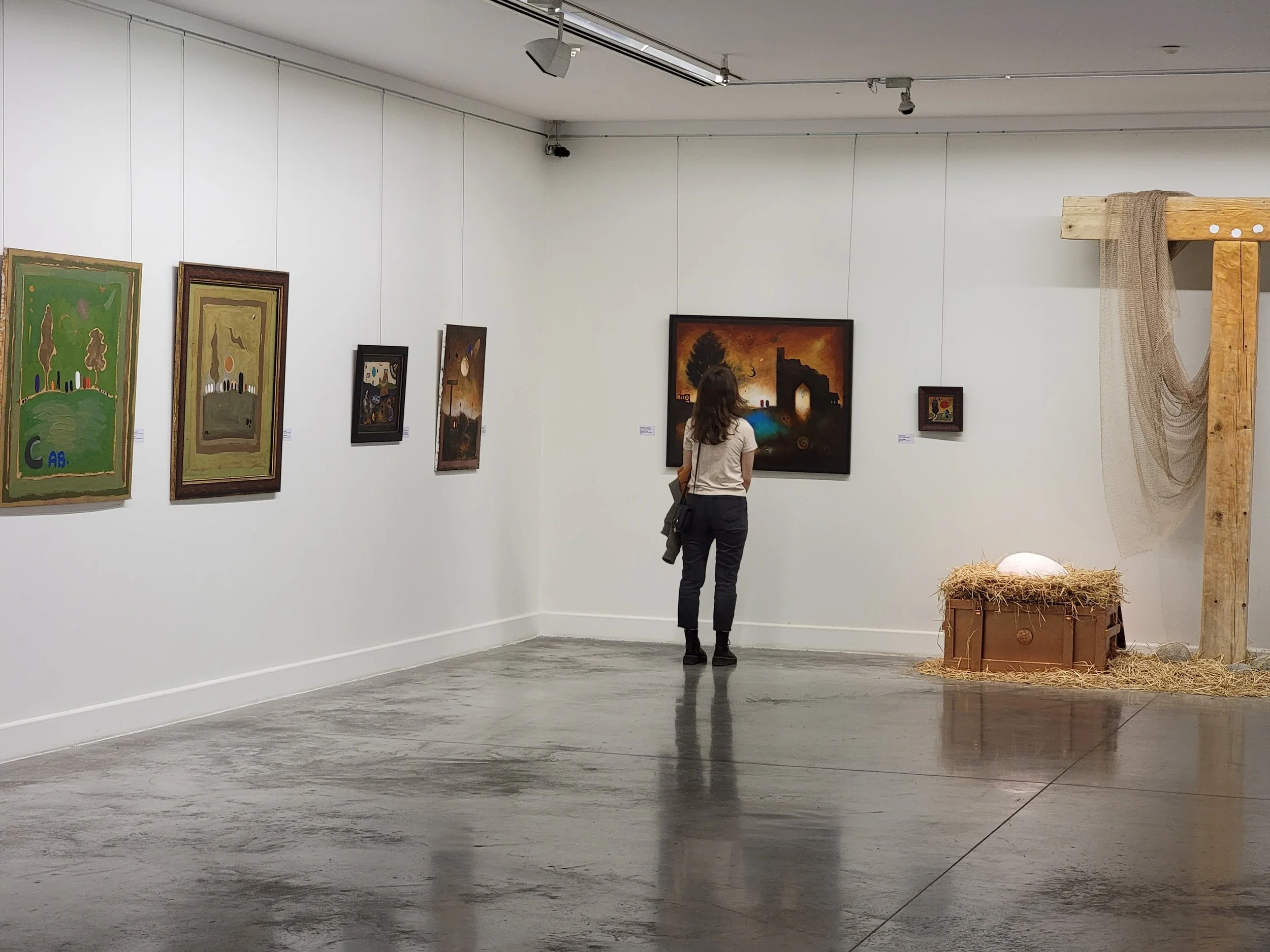 A woman with long brown hair, wearing a white t-shirt and dark pants, is looking at Anton Balanchivadze's paintings on the wall of an georgian nationall gallery. To her right, there is a nativity scene decoration with a chest, hay. 