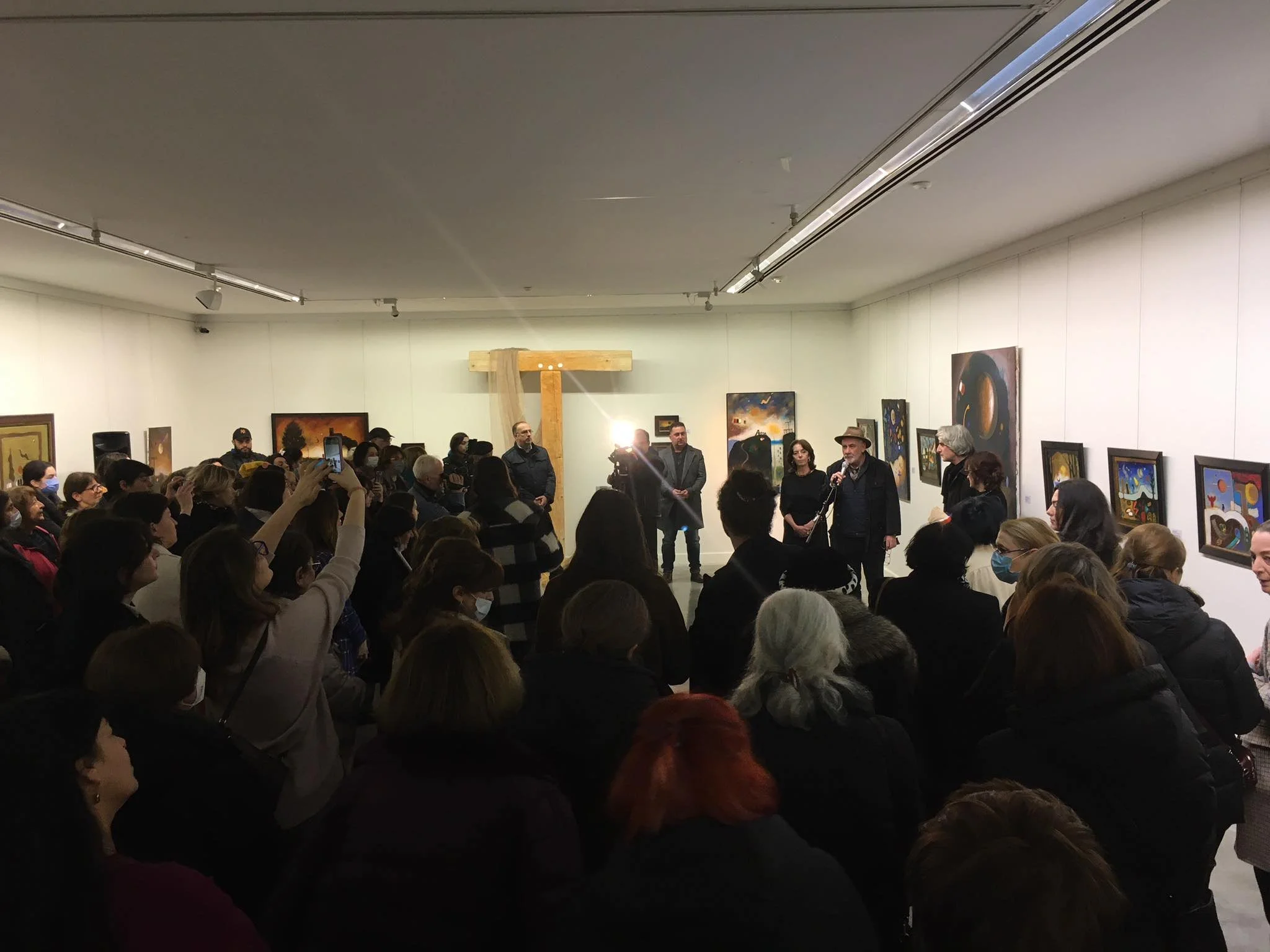 People gathered in an art gallery, listening to a speaker holding a microphone, with paintings displayed on the walls.