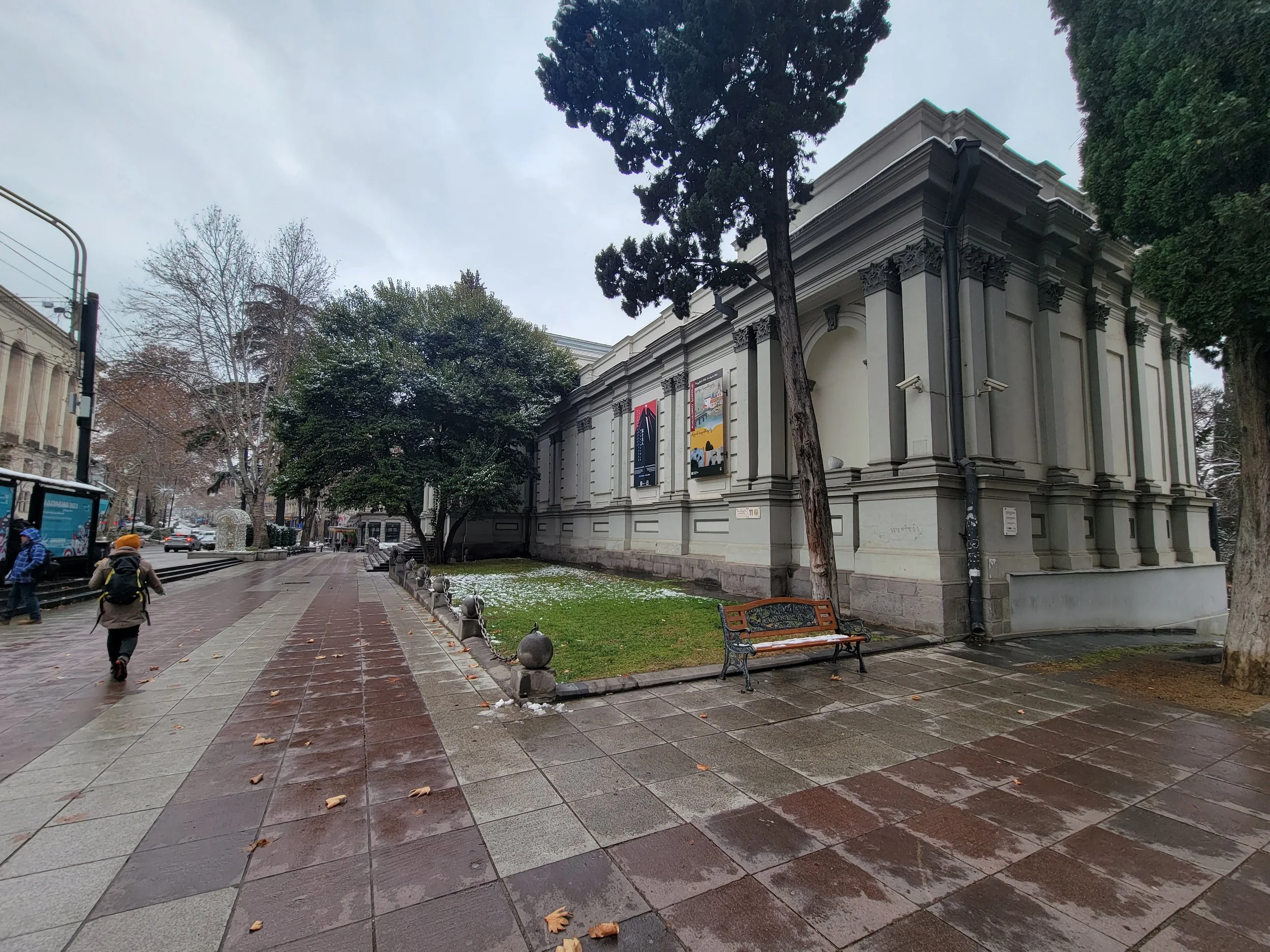 Sidewalk view of a city street on a rainy day with a large historical building on the right, trees lining the sidewalk, a bench, and a few pedestrians walking with umbrellas.
