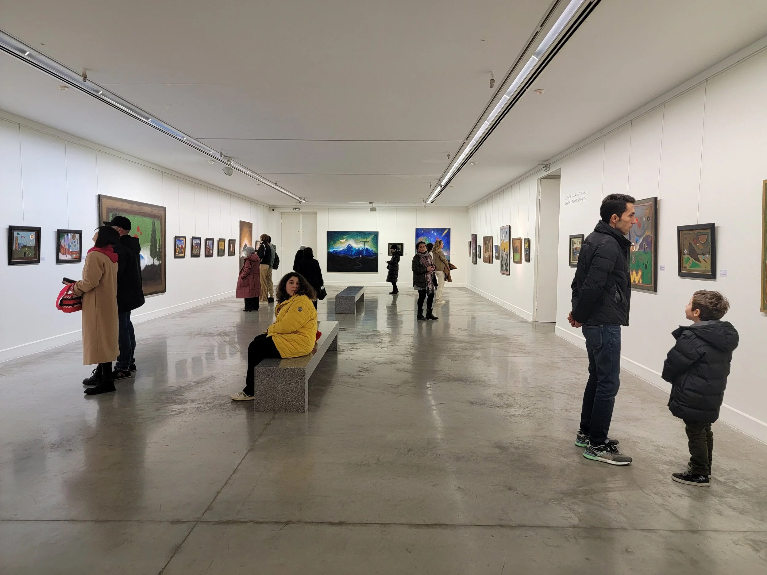 People viewing paintings in an art gallery with white walls and grey polished floor.