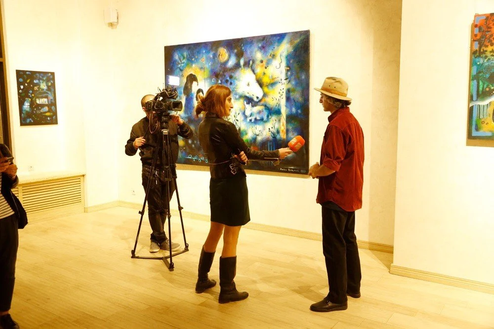 A woman with red hair and black boots conducting an interview with an older man in a hat in an art gallery, with a cameraman filming them and artworks on the wall in the background.