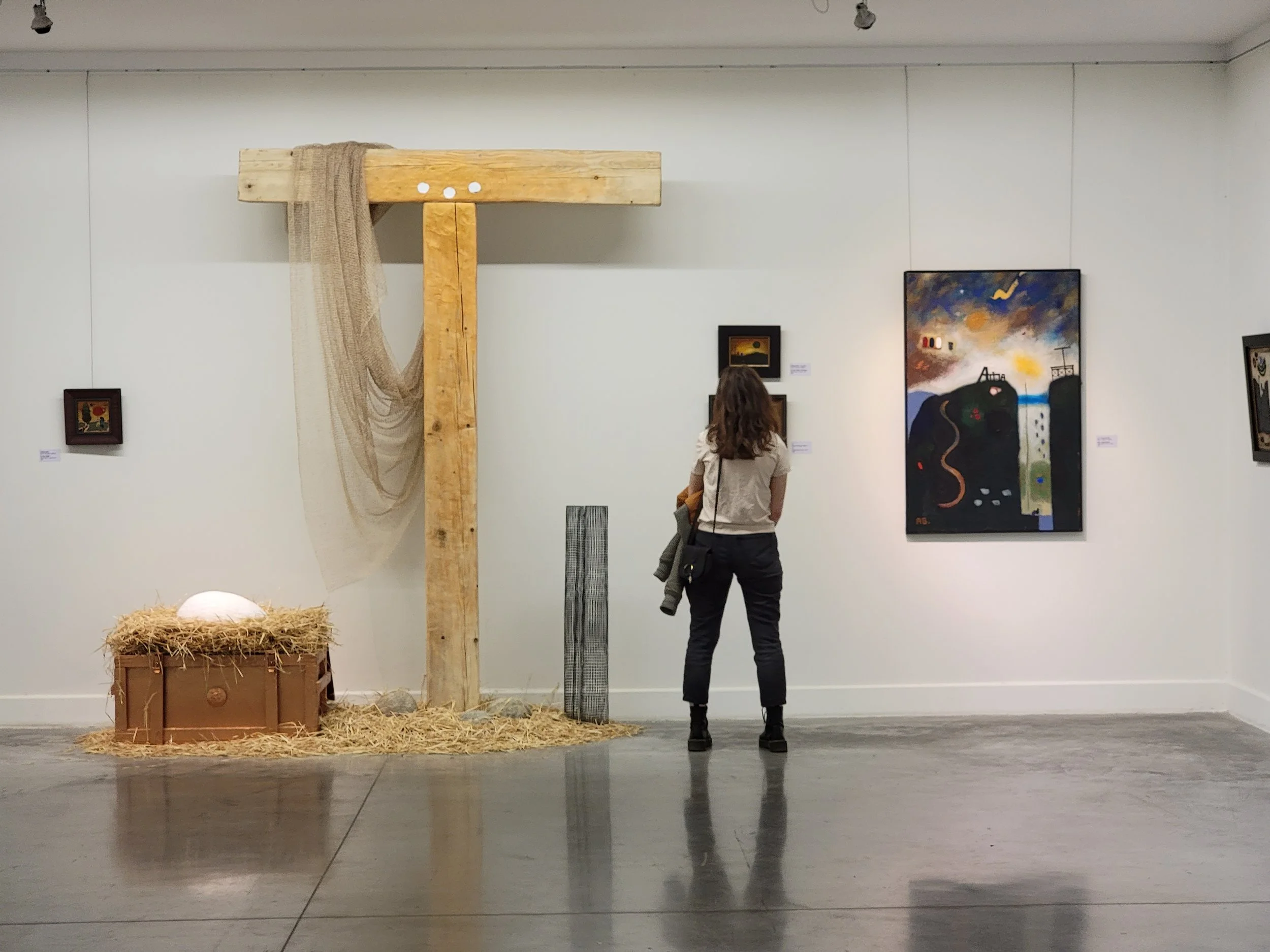 A woman with curly brown hair is in an art gallery, observing paintings on the wall. The gallery has a wooden structure resembling a manger, with a beige fabric draped over it, hay on the floor, and a chest with an egg-shaped object on top. There are