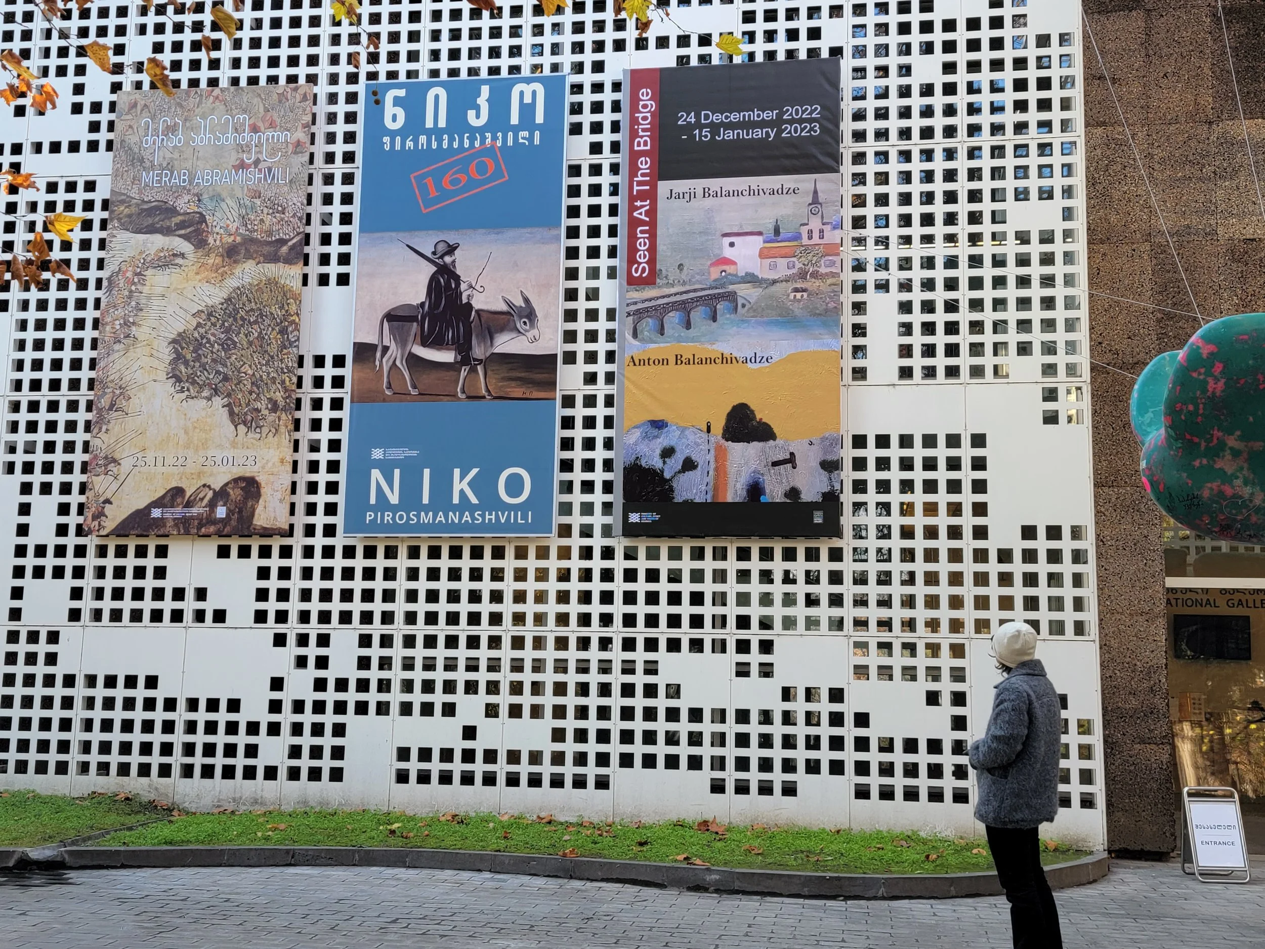 Jarji Balanchivadze and Anton Balanchivadze, Exhibition "Seen at the bridge", woman wearing a gray coat and white hat standing outside in front of georgian national gallery, looking at three large posters advertising art exhibitions.