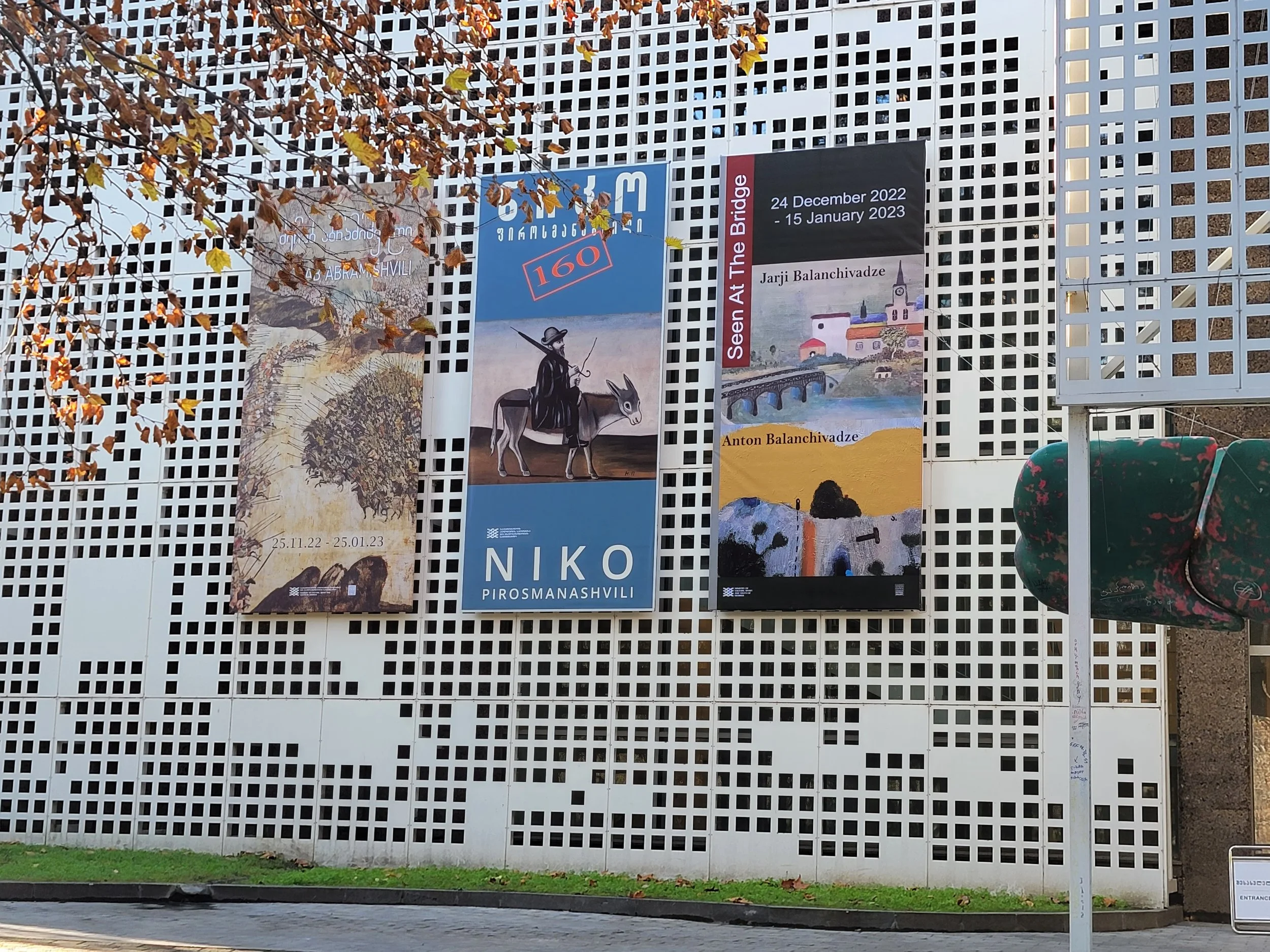 Three vertical banners hanging on a white lattice wall, featuring images and text about art exhibition of Merab Abramishvili, Niko Phirosmani, Jarji and Anton Balanchivadze, with a tree with autumn leaves in the foreground and pedestrian path below. 