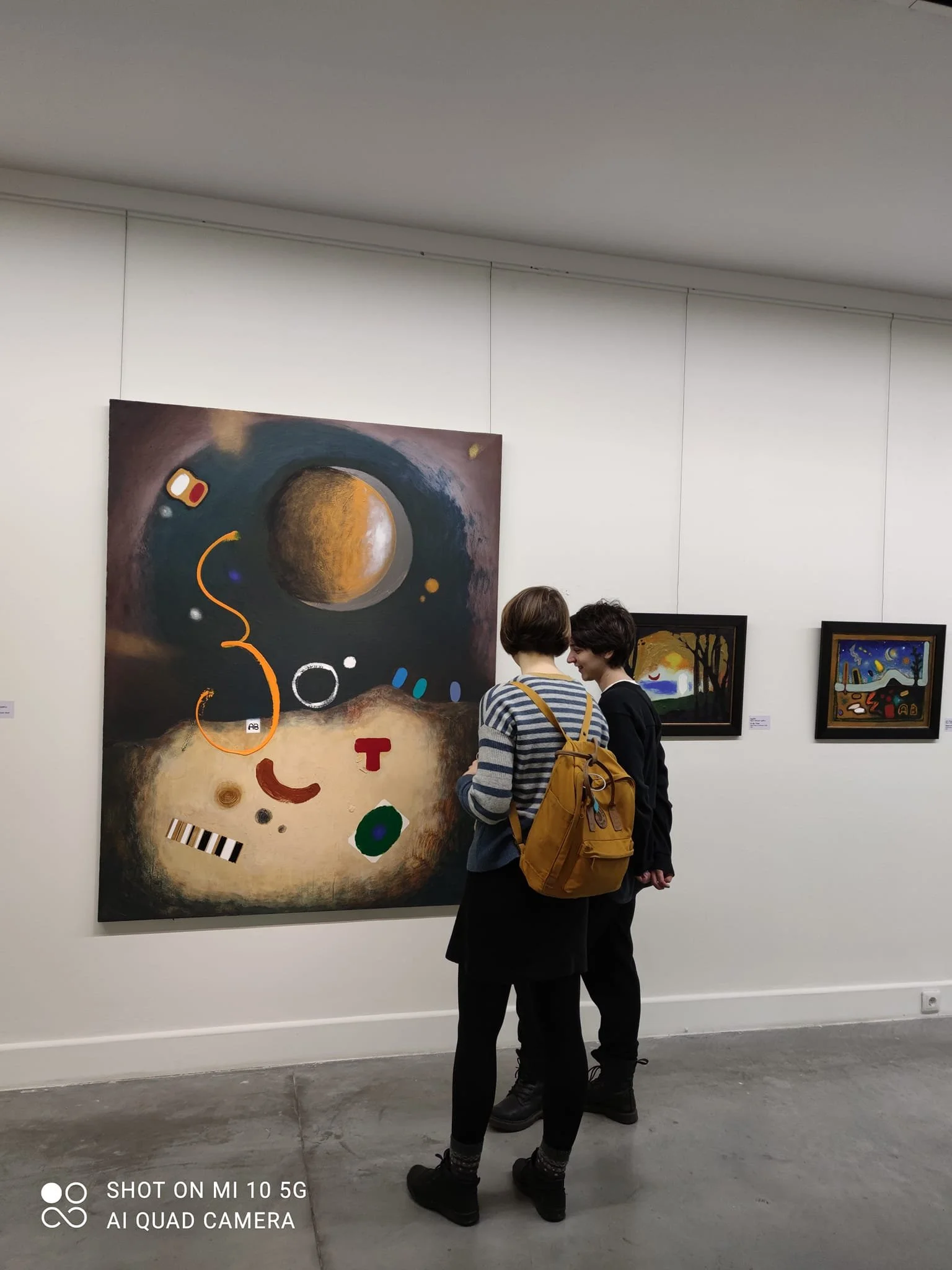 Two people are observing abstract paintings in an art gallery, with one person wearing a yellow backpack and striped sweater, and the other in dark clothing.
