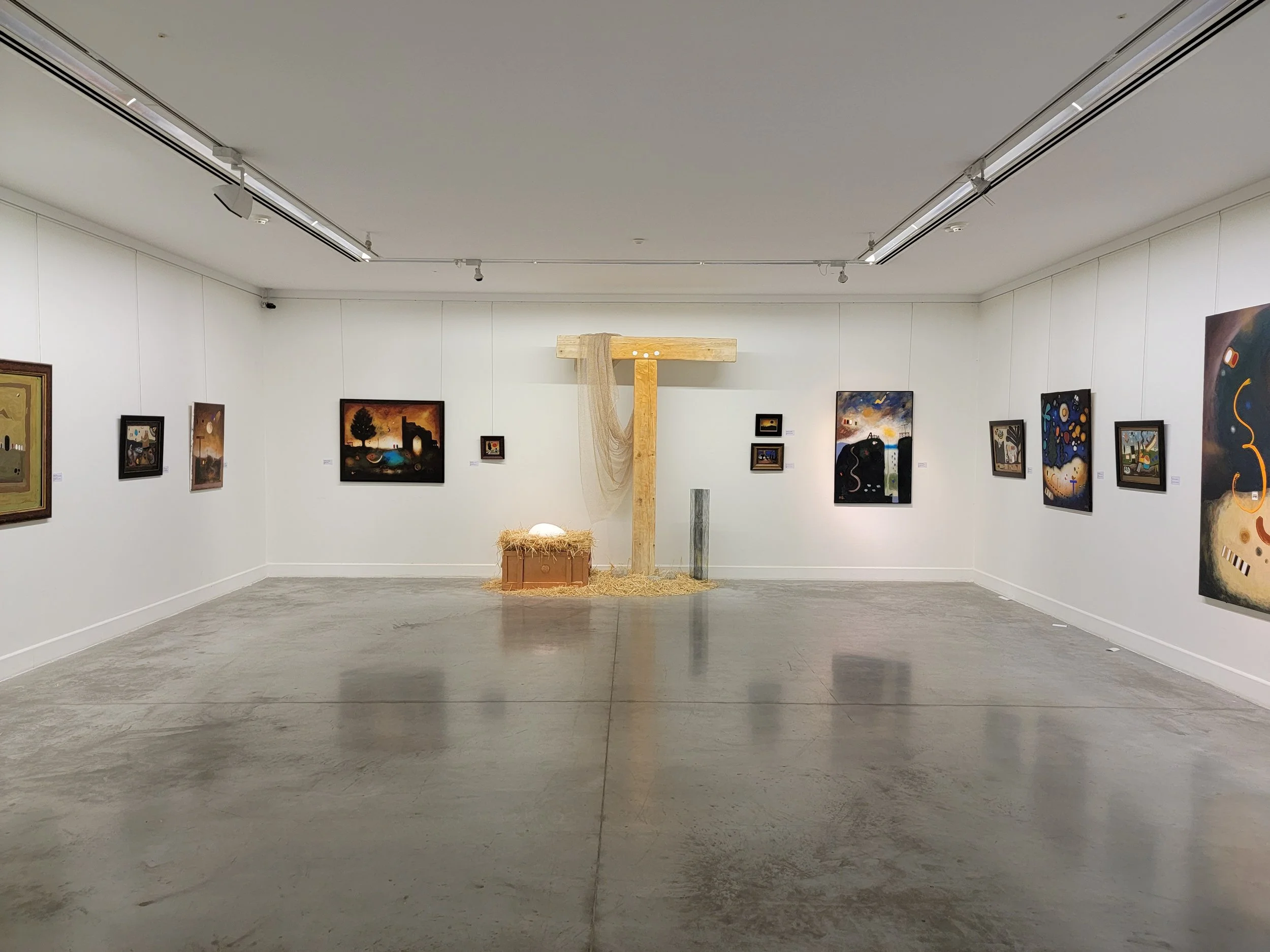Art gallery with white walls displaying various paintings and a wooden cross structure in the center, decorated with hay and a net, with a white egg and straw underneath.